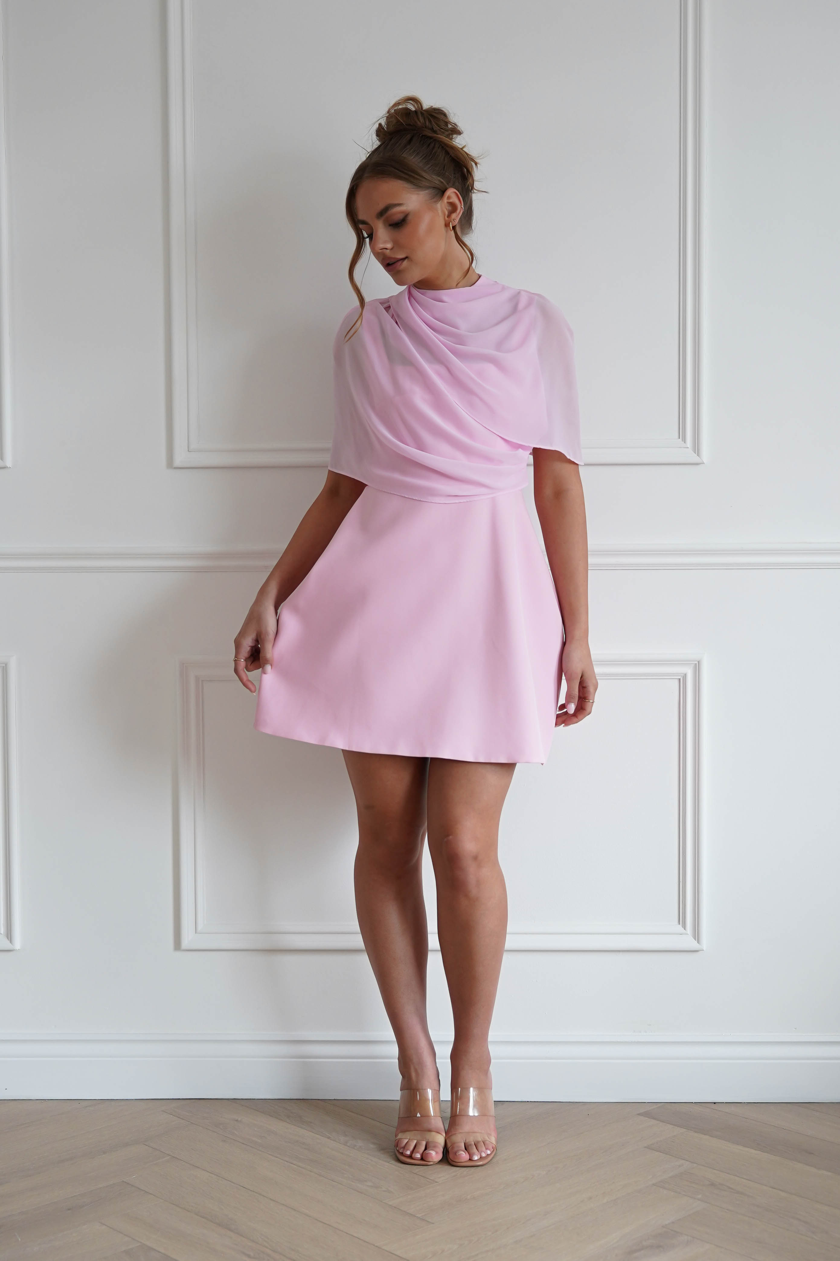Woman wearing a pink mini dress with a sheer chiffon overlay, draped high neckline, flowing cape-style sleeves, and an elegant A-line silhouette, perfect for weddings, baby showers, engagement parties, and special occasions.