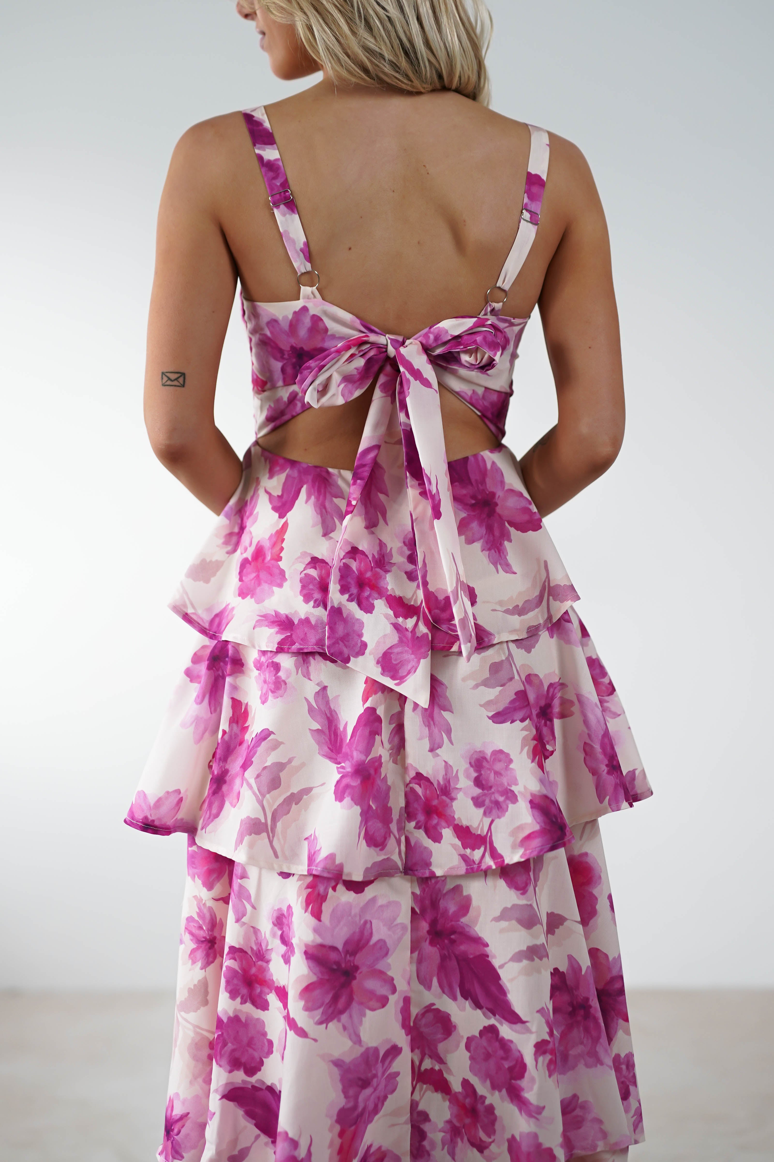 A person wearing a floral print dress adorned with pink and purple flowers stands with their back to the camera. The wedding guest attire features a bow and layered ruffles, complemented by adjustable straps, set against a plain background. Light hair cascades gently down their back.