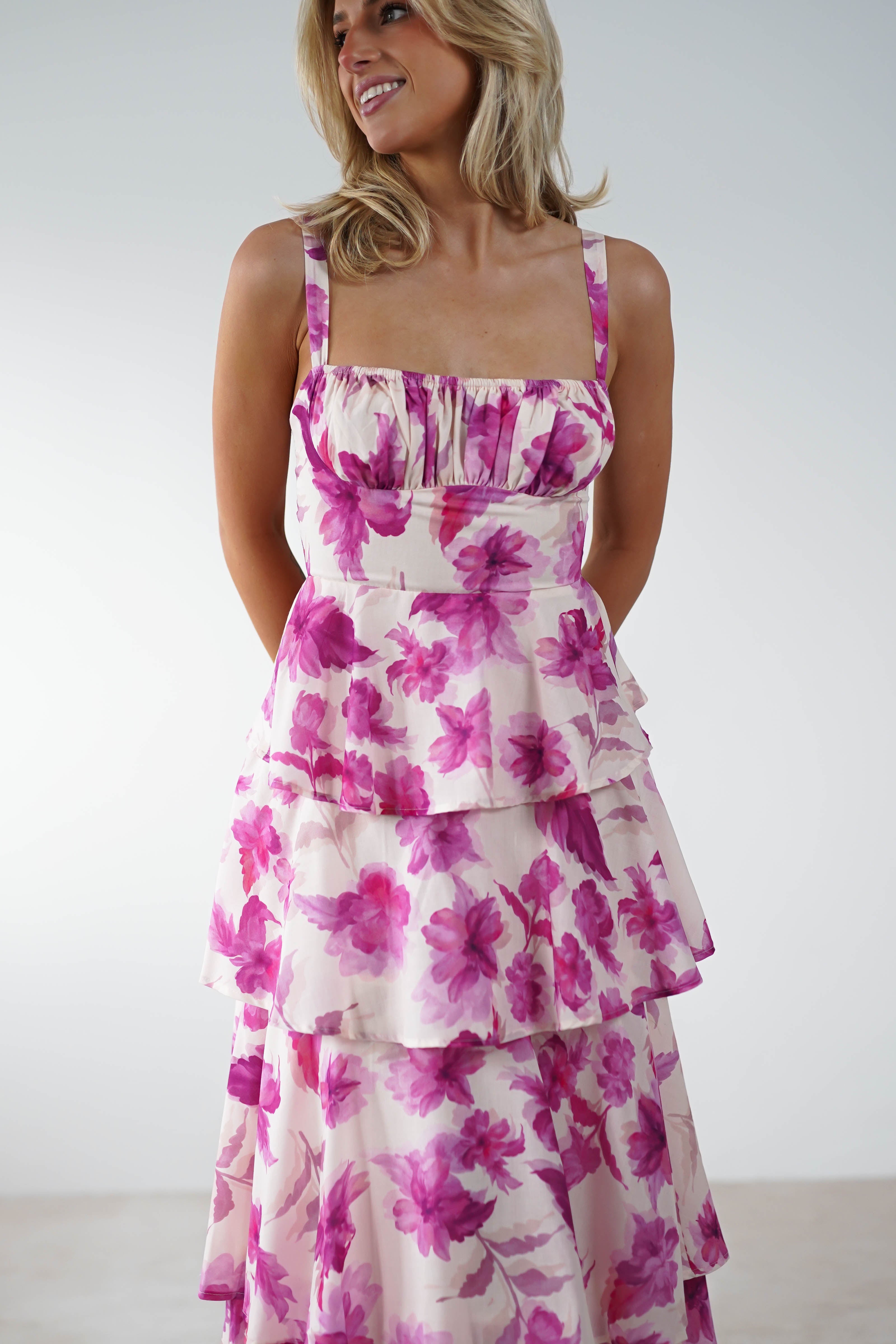 A woman in a sleeveless dress with pink and purple floral patterns smiles while looking to the side. Perfect for a wedding guest, the dress features layered tiers, adjustable straps, and a fitted bodice. She has blonde hair and stands against a plain light background.