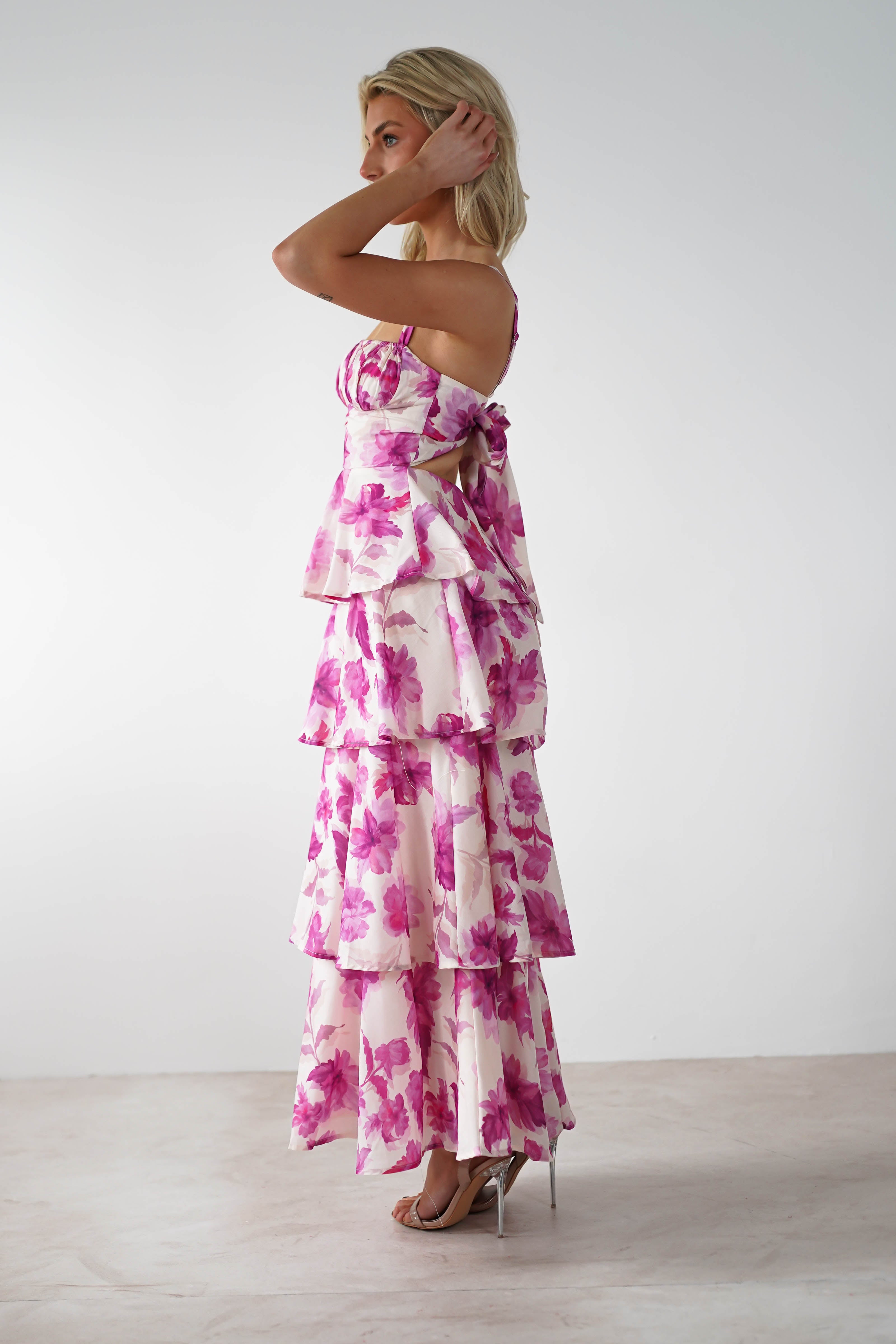 A woman in a tiered floral dress with shades of pink and purple, perfect for a wedding guest, stands against a plain white background. She is turned to her left, with hands near her face, showcasing the multiple ruffles and adjustable straps of her open-back attire.