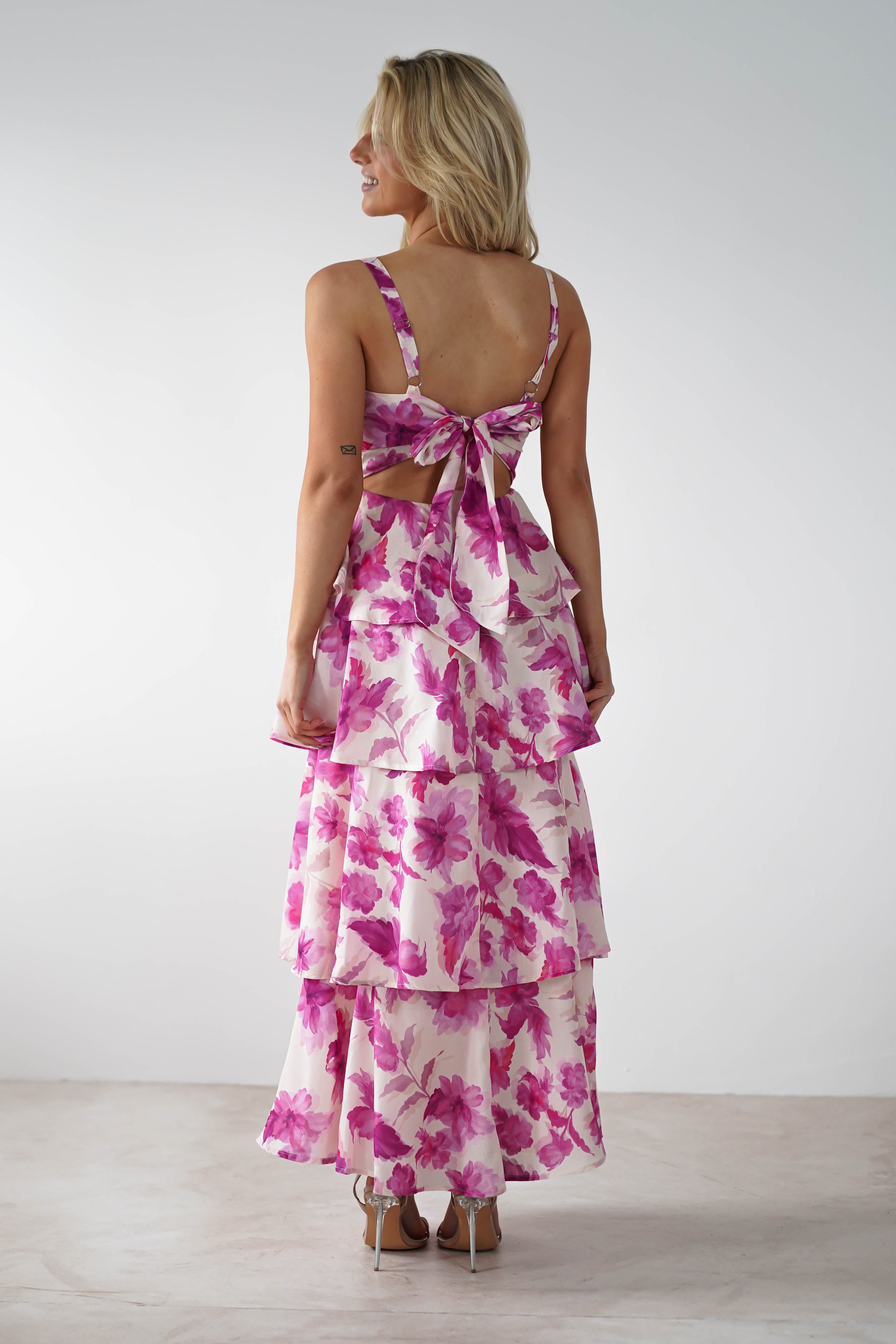 A woman with blonde hair stands with her back to the camera, wearing a sleeveless dress adorned in a pink floral print. Perfect for a wedding guest, the dress features tiered layers and adjustable straps, complete with a large bow in the minimalistic indoor setting.