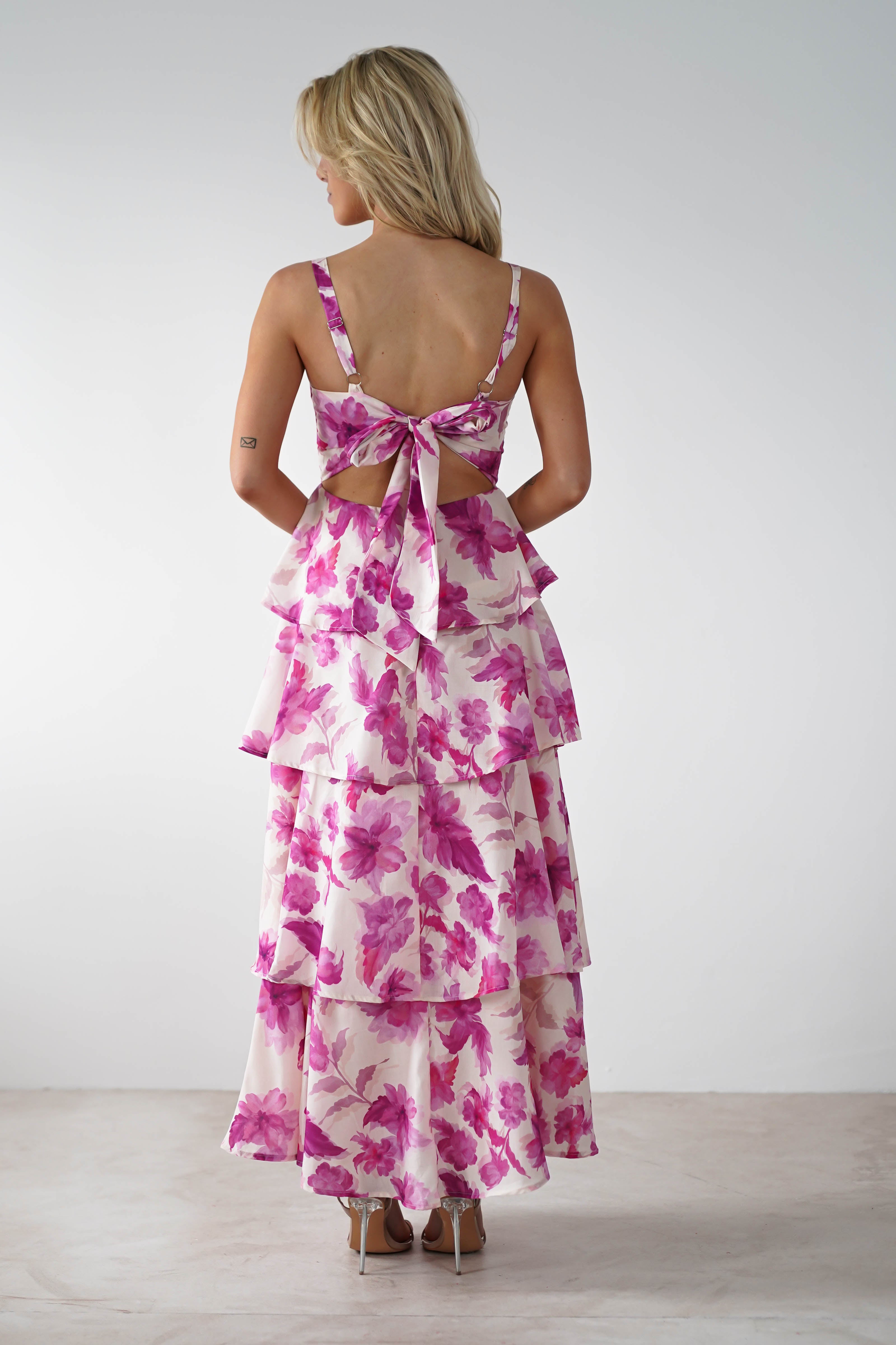 A person with blonde hair stands with their back to the camera, wearing a sleeveless, floor-length, tiered dress adorned in a pink floral print. Designed for a wedding guest, this elegant ensemble features adjustable straps and a charming bow at the back against a neutral backdrop.