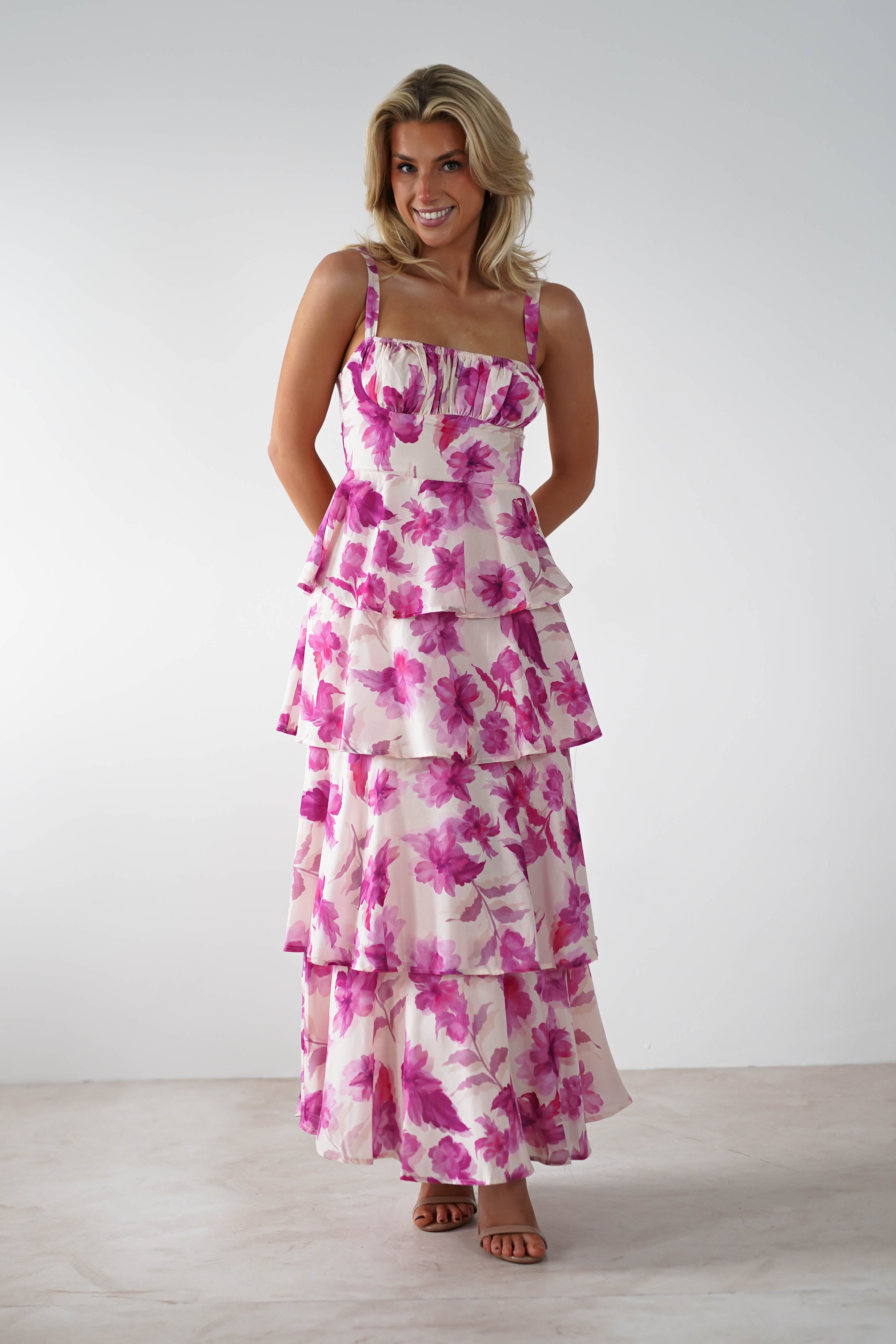 A smiling woman stands against a plain background, wearing a tiered, sleeveless dress with beautiful floral print and adjustable straps. She has her hands clasped in front of her and is wearing sandals—an ideal choice for a wedding guest.