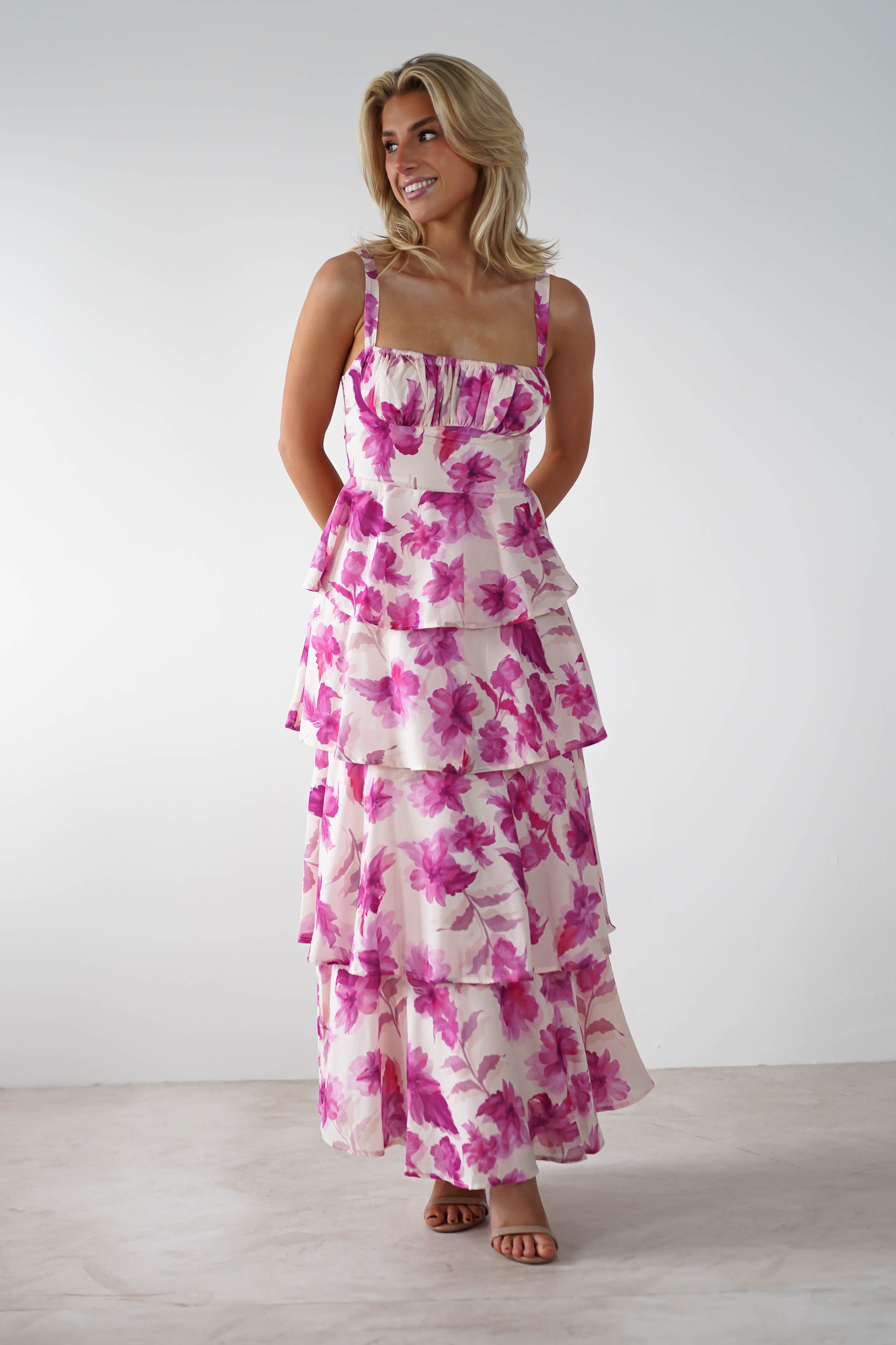 A person wearing a sleeveless, tiered ruffle dress with adjustable straps and a pink floral print stands against a plain white background. The floor-length dress is perfect for a wedding guest, and they are smiling with hands clasped behind their back.