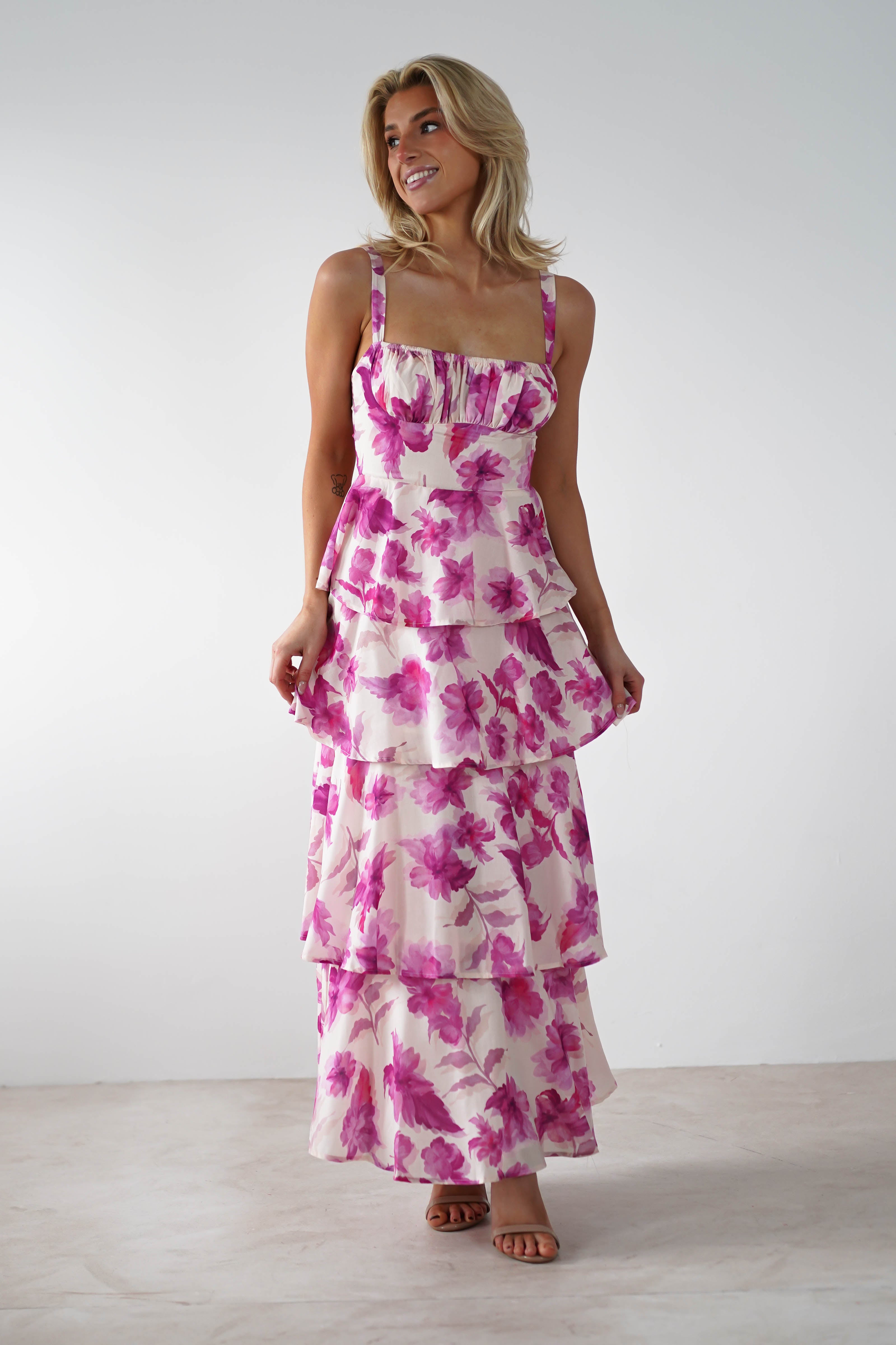 A person stands wearing a tiered, pink floral dress with adjustable straps. They have shoulder-length blond hair and are smiling in a bright, minimalistic room with a white background—perfect attire for a charming wedding guest.