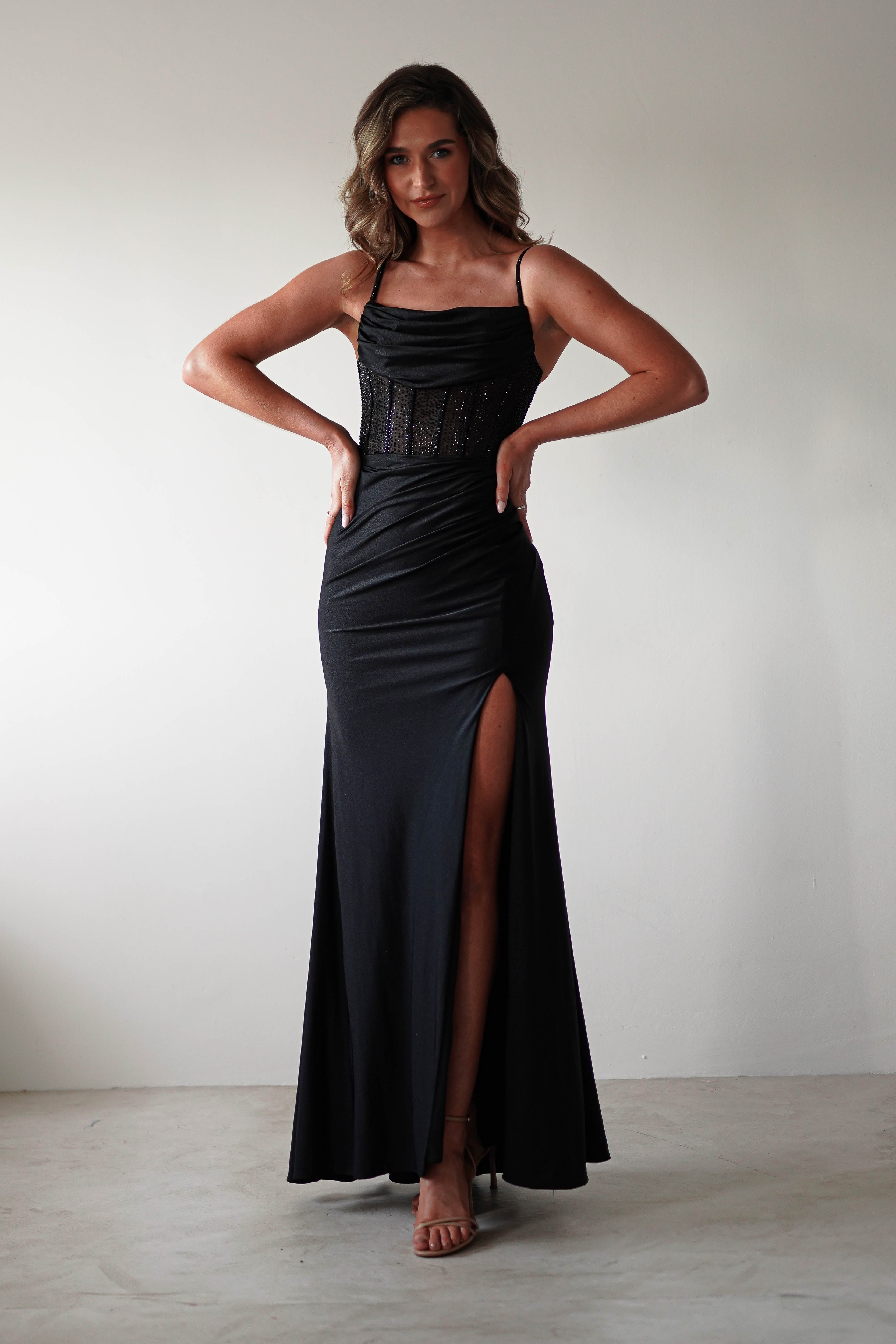 A woman stands confidently against a plain white background, hands on hips. She wears a stunning, fitted gown with a long black satin skirt and high slit that complements the sheer rhinestone bodice perfectly.