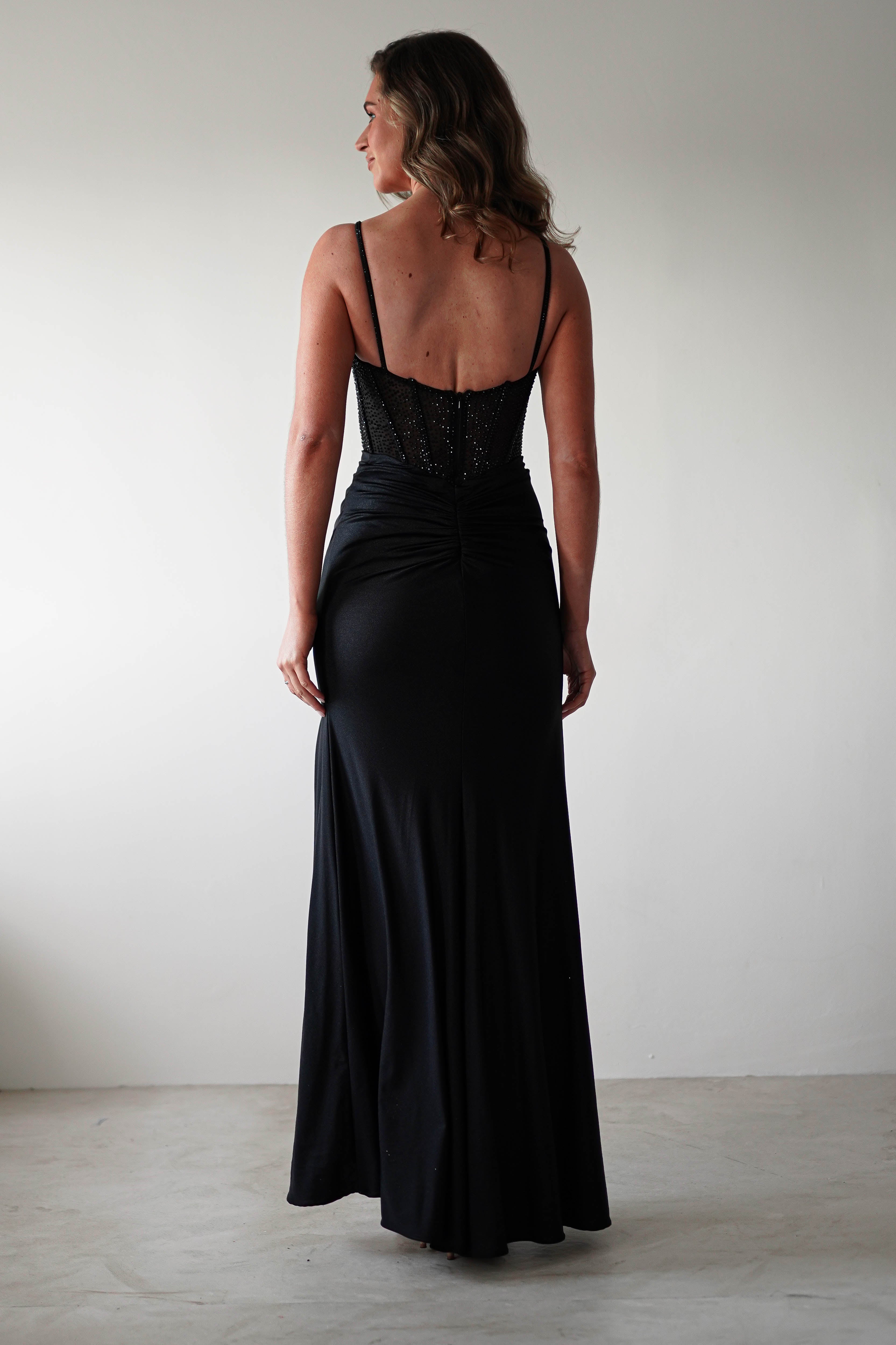 A woman in a black, floor-length evening gown with thin straps stands facing away. The fitted gown boasts a sheer rhinestone bodice and a flowing satin skirt. The background is a plain white wall, and the woman has shoulder-length brown hair.