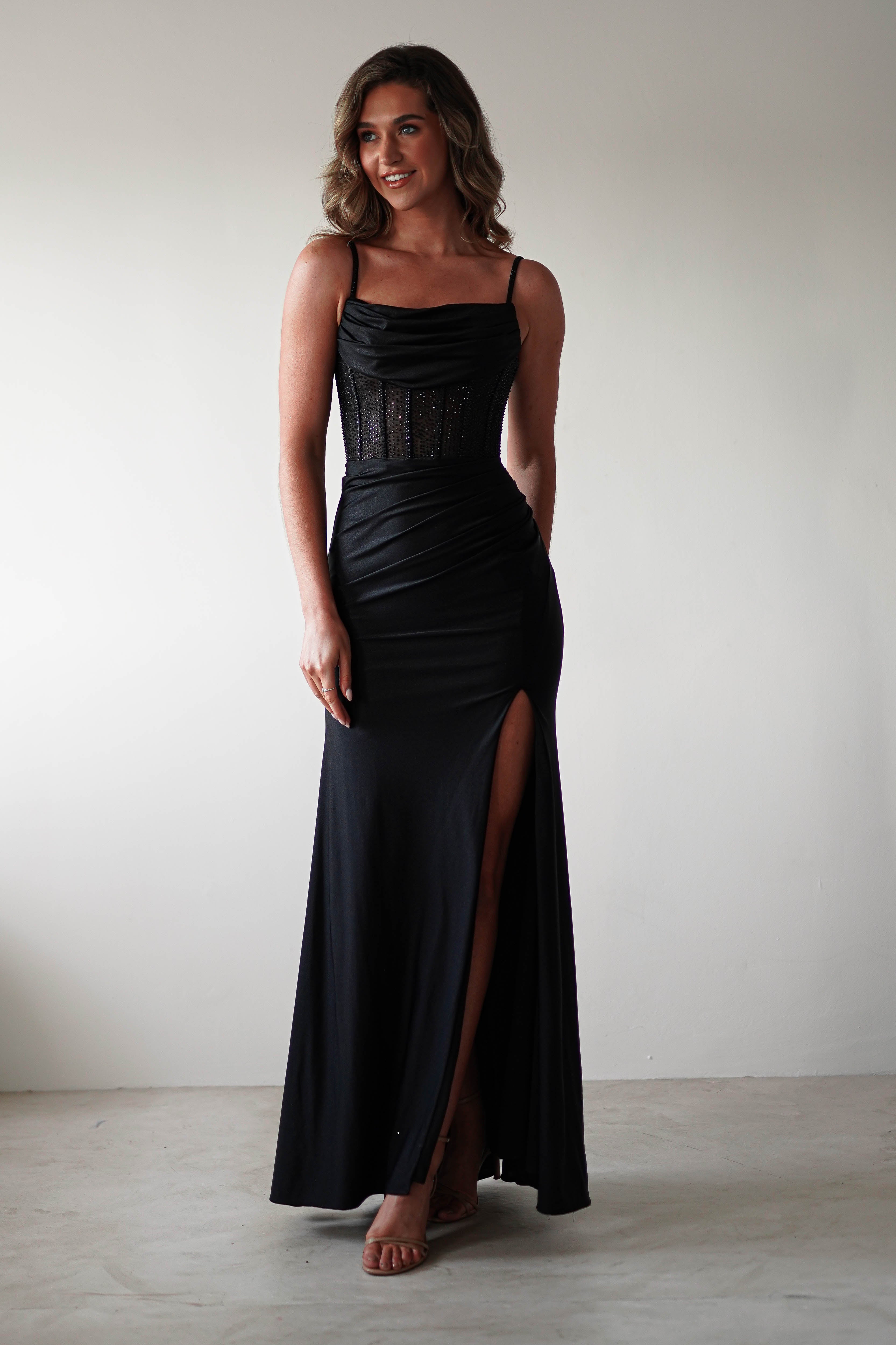 A person in a black, sleeveless evening gown with a sheer rhinestone bodice stands against a plain backdrop. The fitted dress features a satin skirt with a high slit. They have shoulder-length wavy hair and are wearing nude heels.