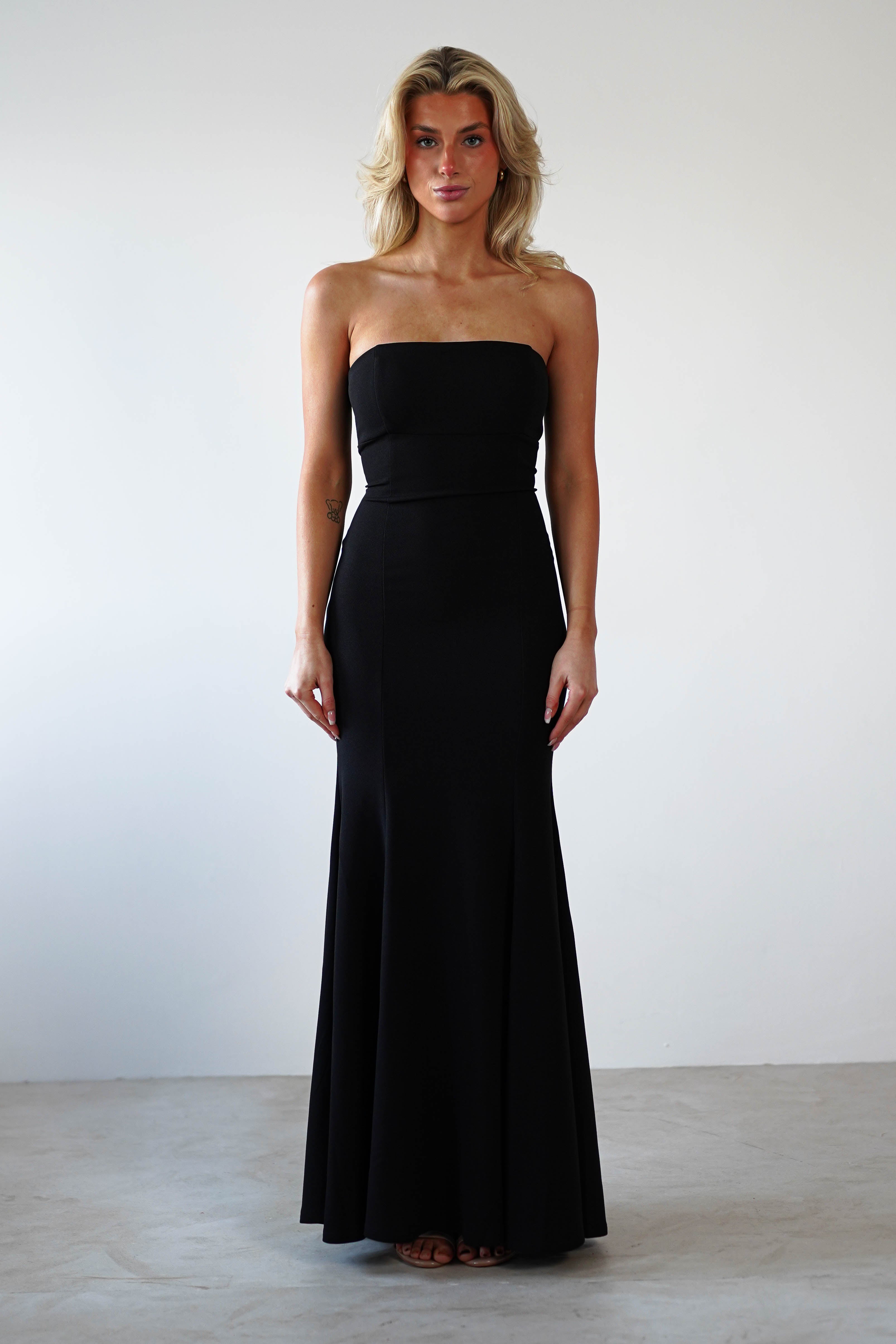 A woman in a strapless black evening gown stands against a plain white background. The formal occasion wear features a fitted bodice and flared mermaid skirt, perfectly complementing her shoulder-length blonde hair.