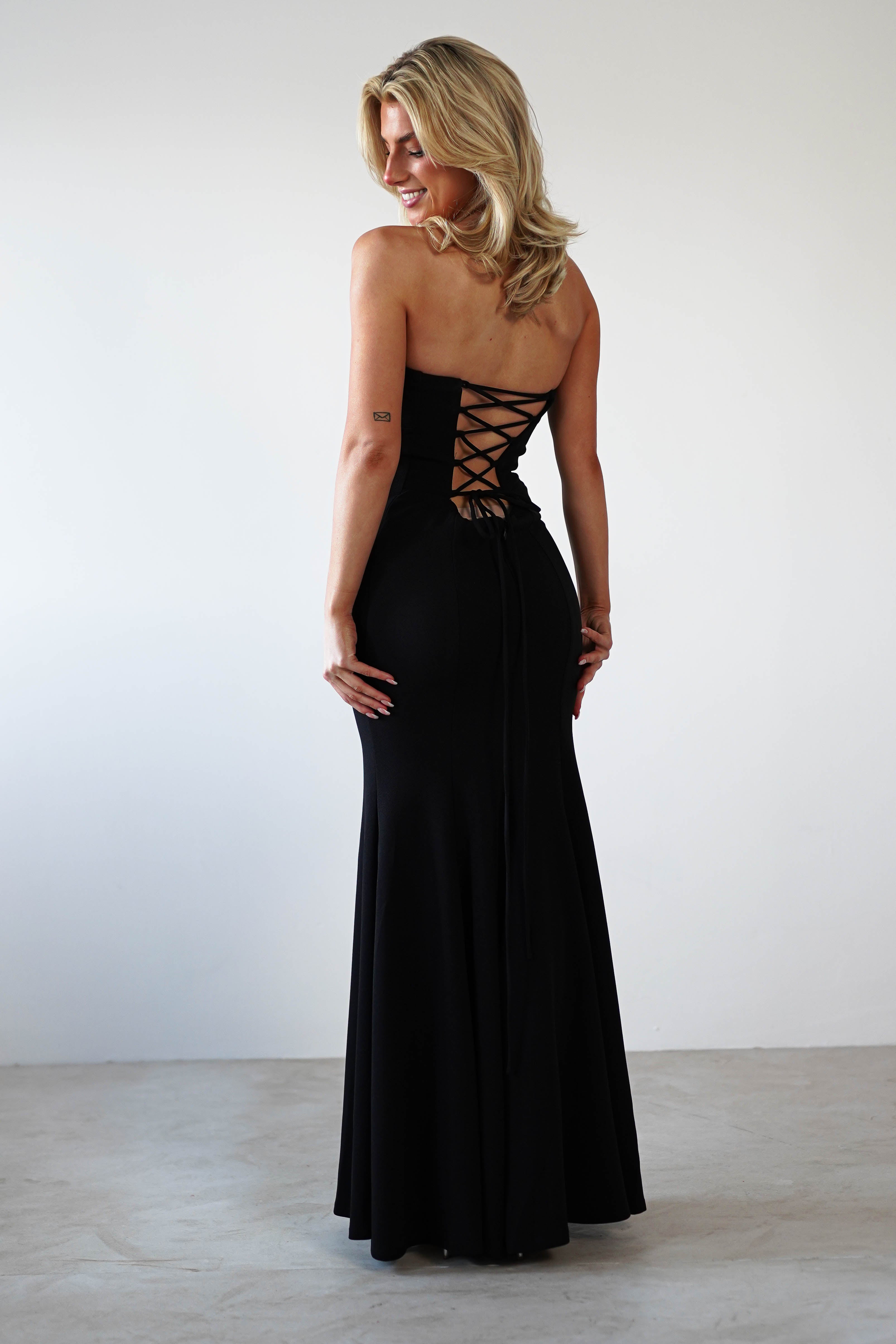 A person wearing a stunning black evening gown with a lace-up back stands against a plain white background. The strapless mermaid dress graces their figure elegantly as they turn slightly, smiling, with blonde hair and a small tattoo on their upper arm.