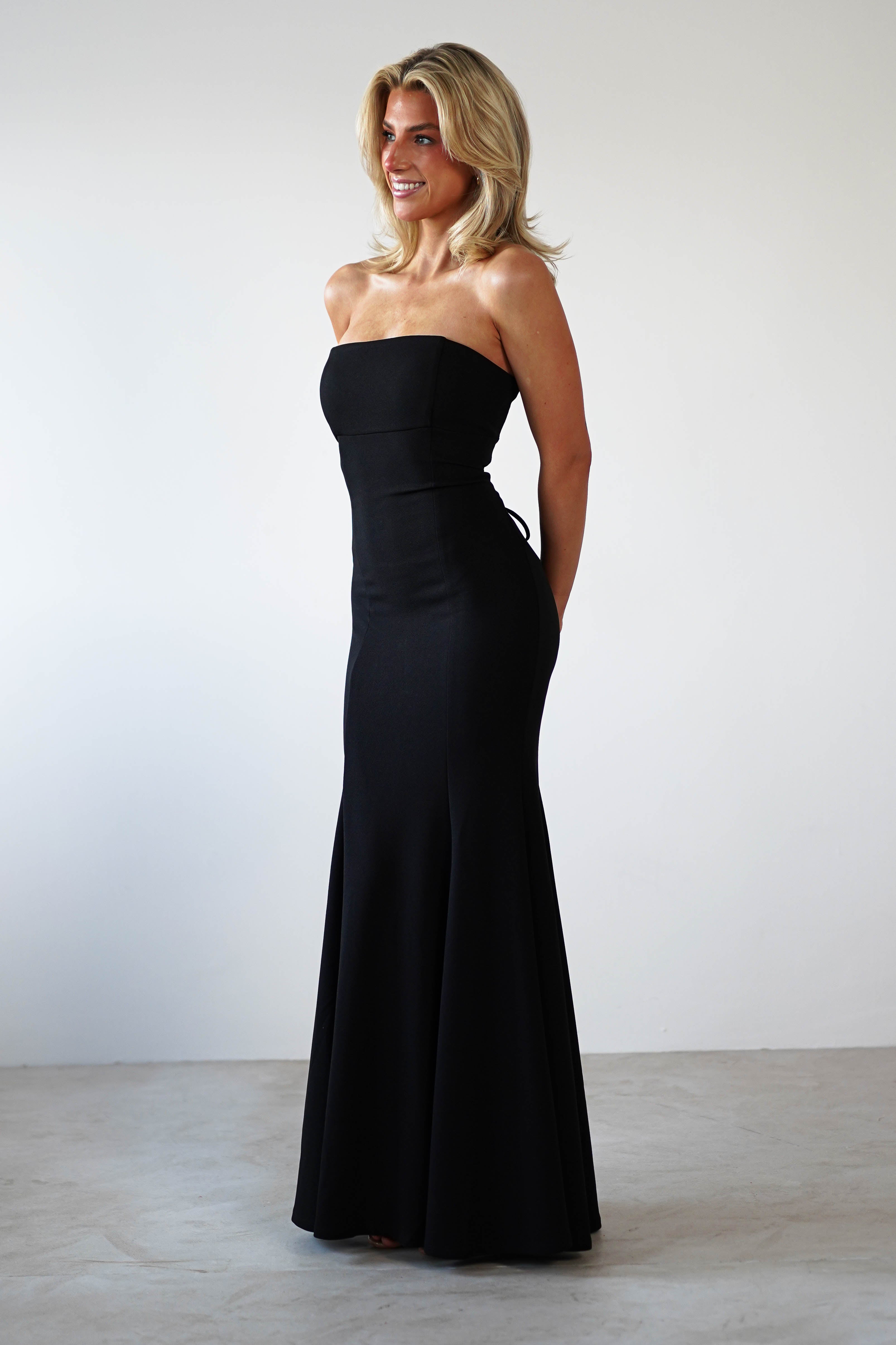 A woman in a sleek, strapless mermaid dress stands smiling with her hands behind her back. She has shoulder-length blonde hair and is set against a plain white background, exuding elegance perfect for any formal occasion.