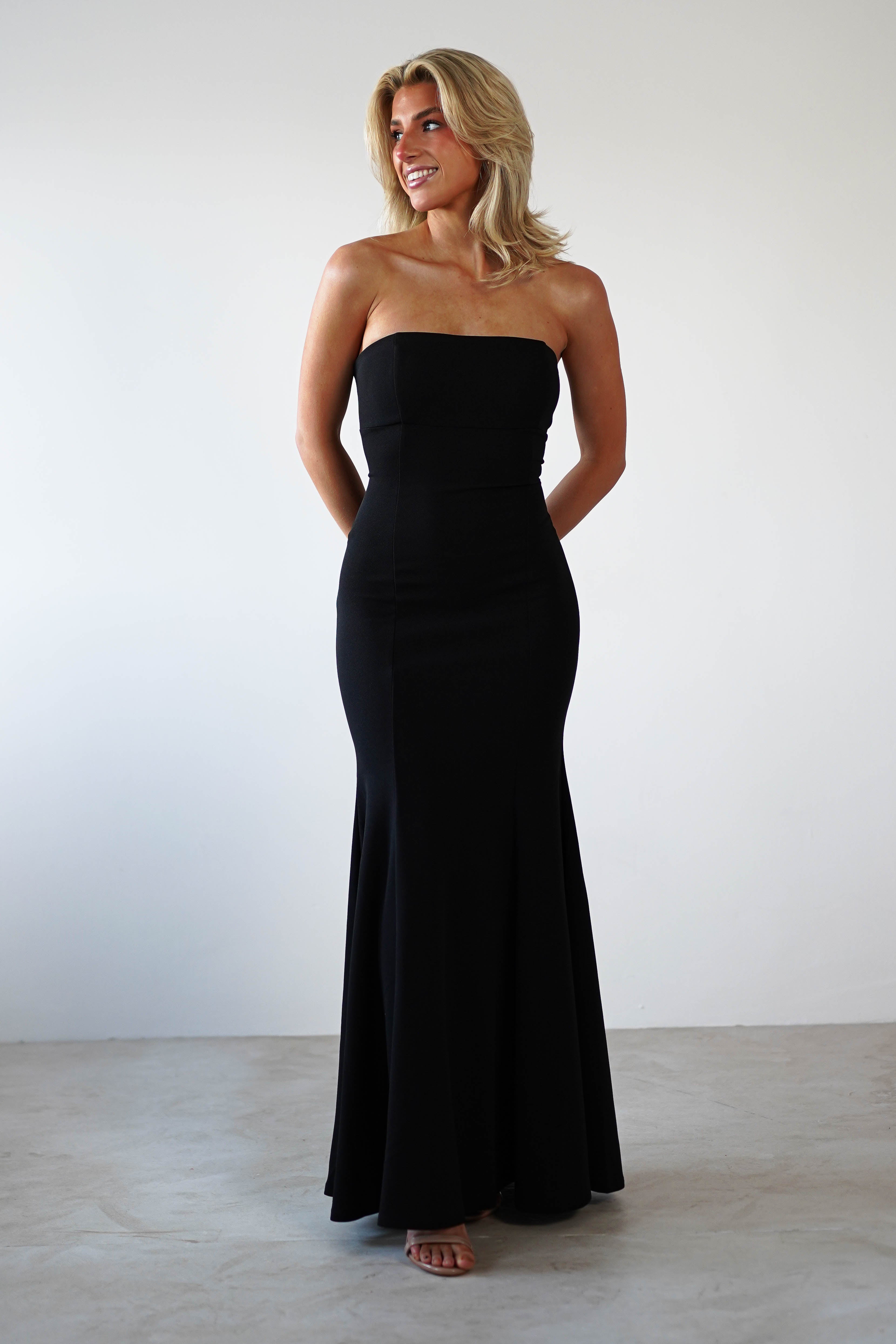 A person elegantly dons a strapless mermaid dress, standing against a plain white background. With shoulder-length blonde hair, they are smiling warmly, hands tucked behind their back—perfectly embodying formal occasion wear in a stunning black evening gown.