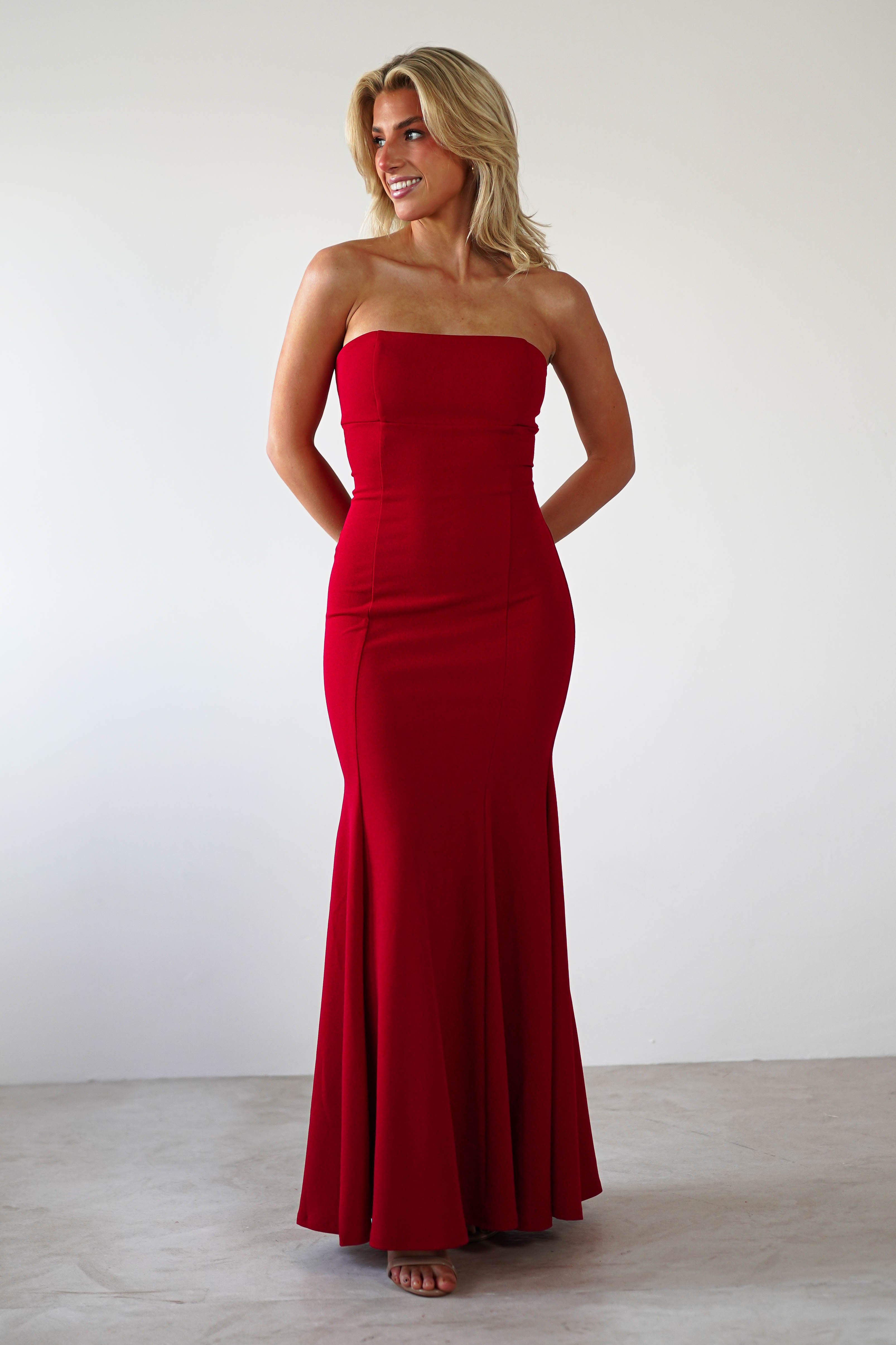 A woman in a strapless evening gown with a striking mermaid silhouette stands against a plain, light background. She smiles, gazing slightly to the side. The floor-length red dress is fitted with a flared bottom, perfect for formal events, as her hands rest gracefully behind her back.