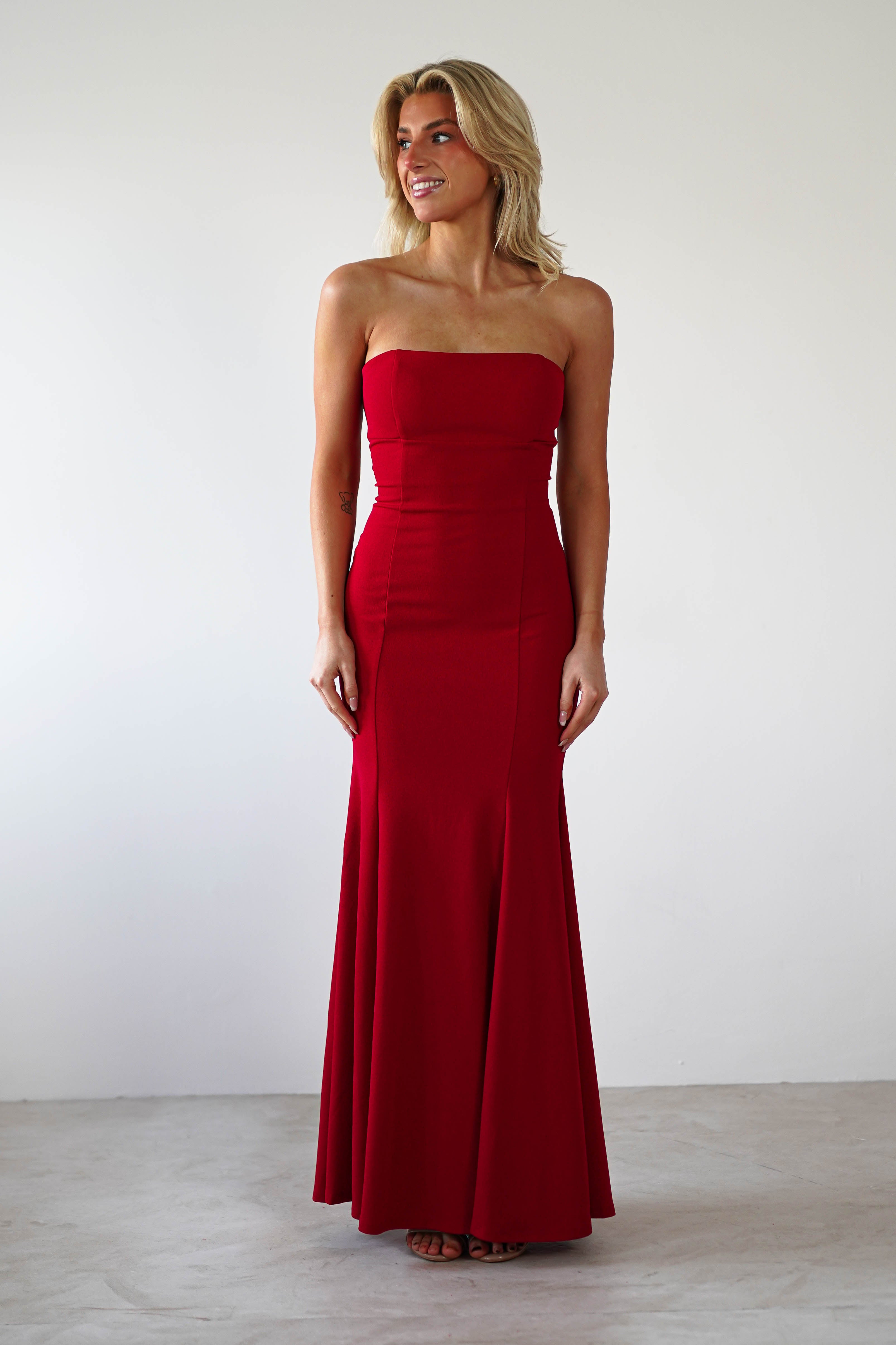 A woman wearing a strapless evening gown stands against a plain white background, exuding elegance. The floor-length red dress, perfect for formal events, highlights her smile as she looks to her right.