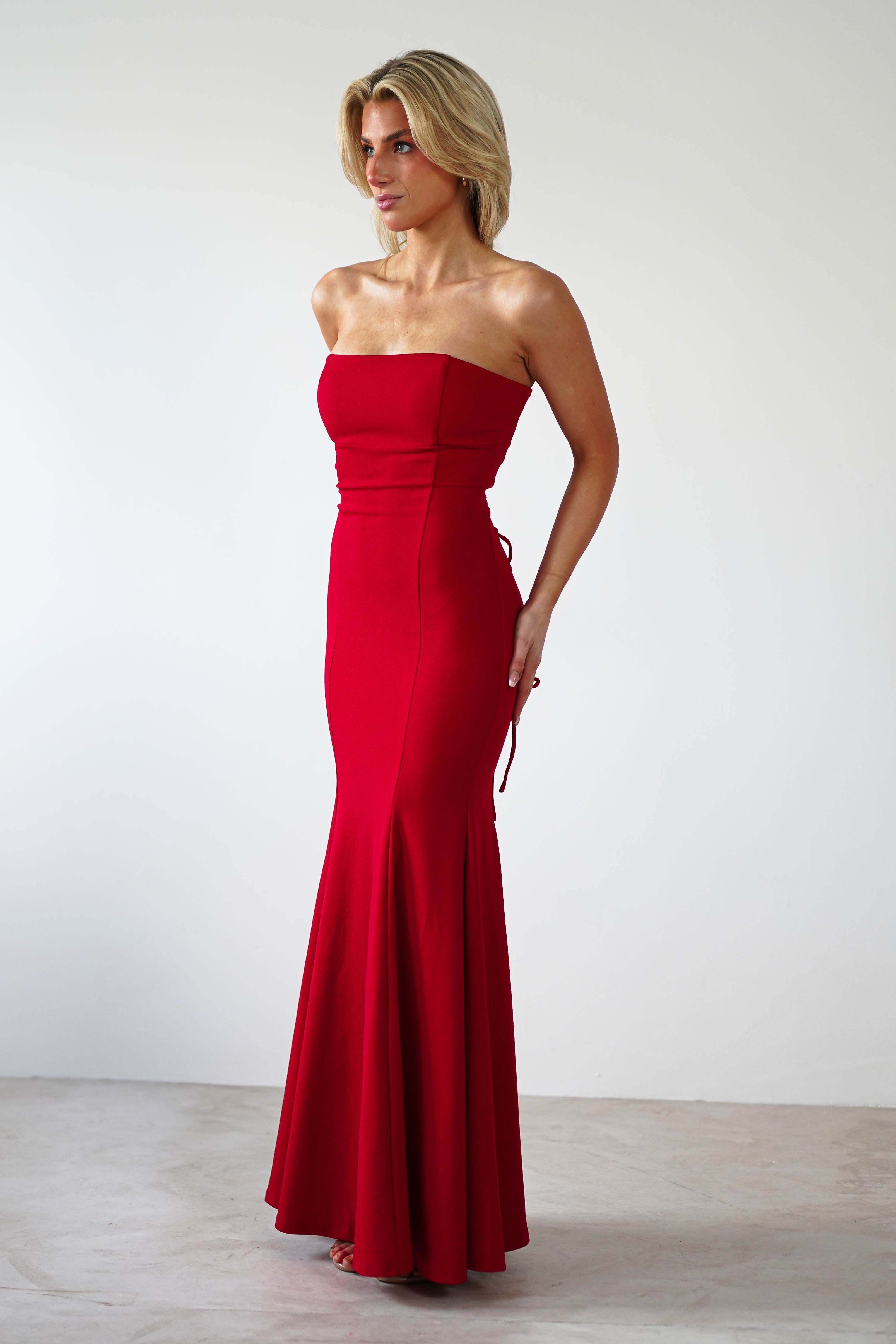 A person wearing a red strapless evening gown with a mermaid silhouette stands against a plain white background. The dress is fitted at the top and flares slightly at the bottom. The individual, with short, light hair, gazes to the side, exuding elegance ideal for formal events.