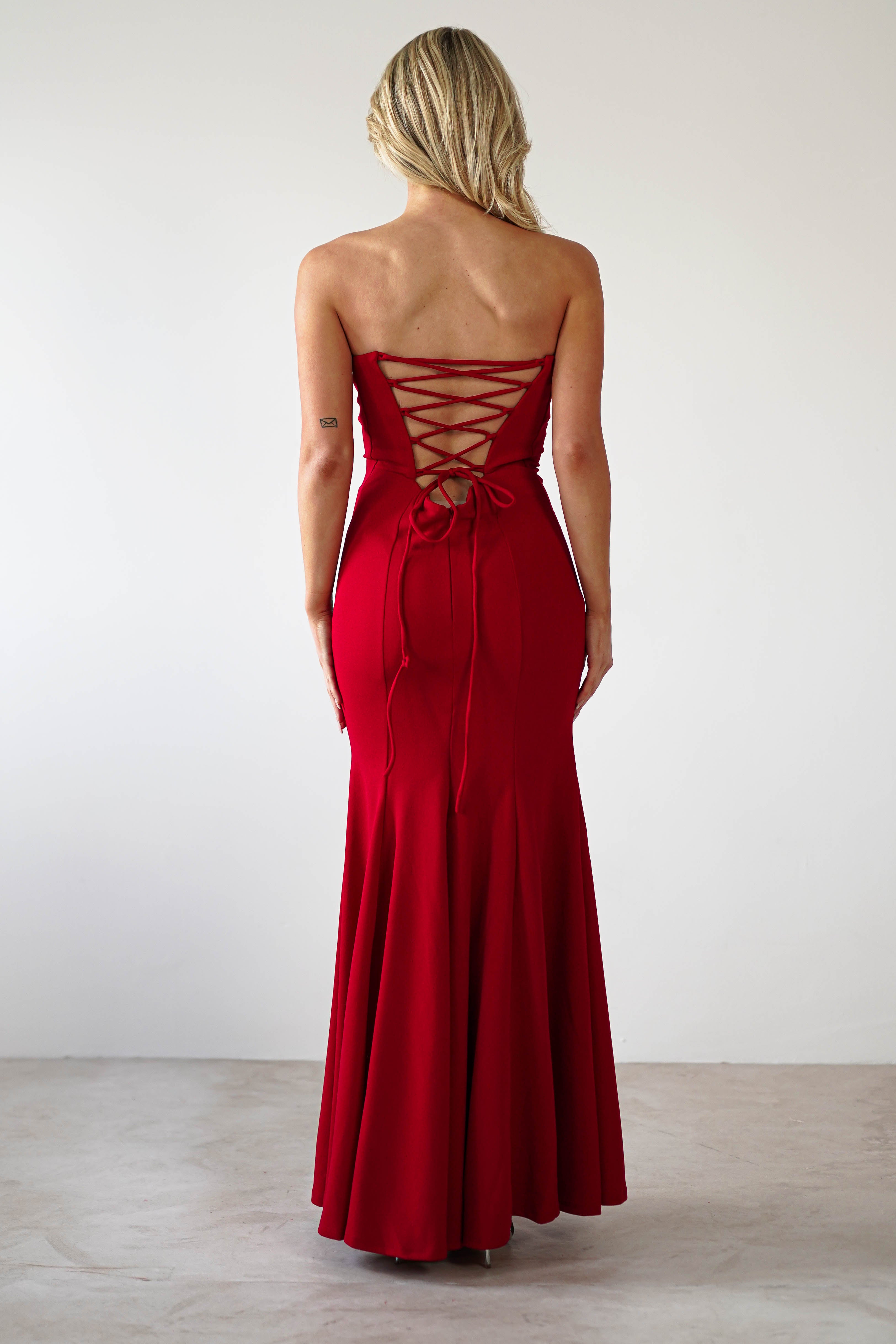 A rear view reveals a person in a strapless, floor-length red dress featuring a corset-style lace-up back and mermaid silhouette. Their long blonde hair cascades as they stand gracefully, ready for formal events in this stunning evening gown.