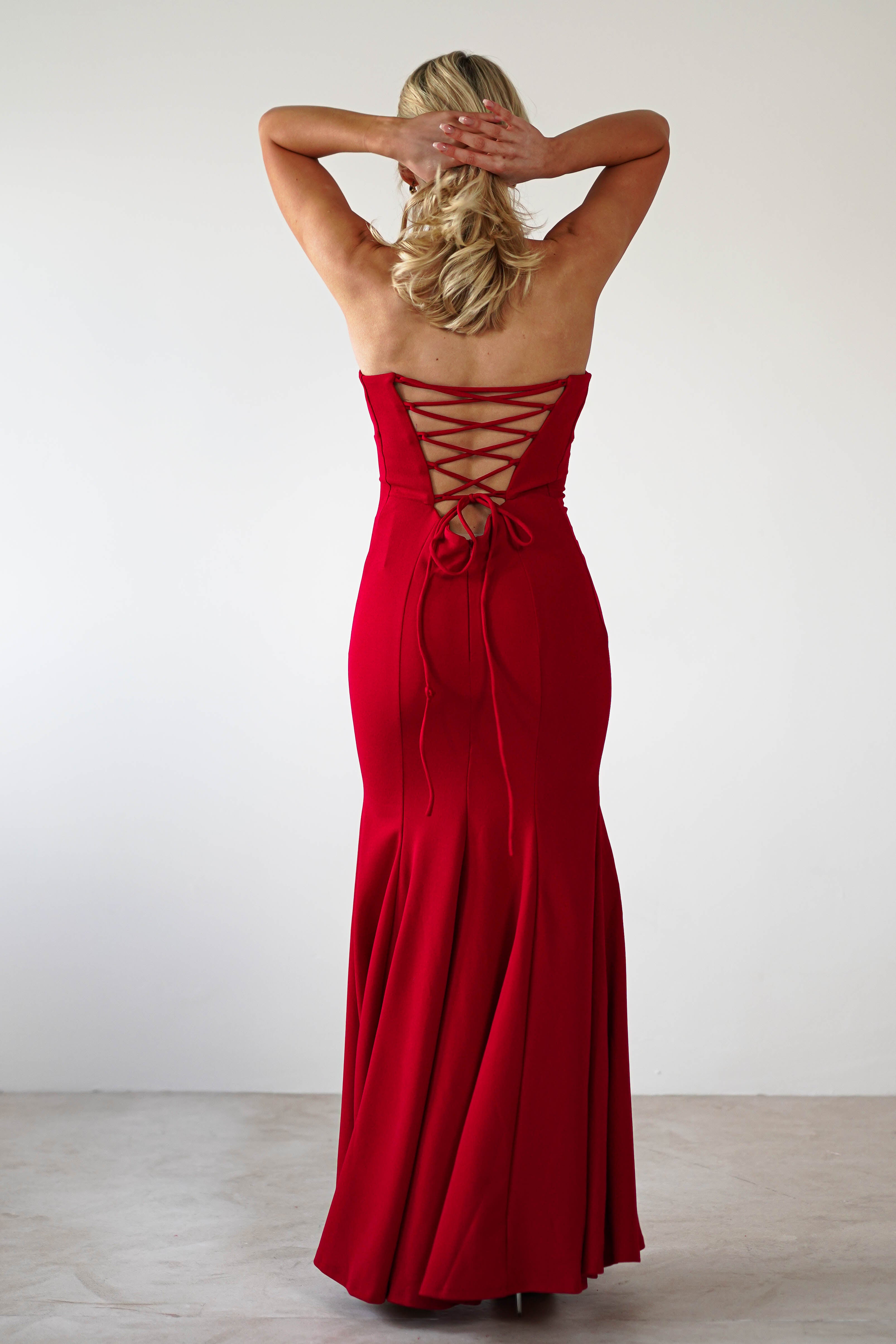 A person with blonde hair poses in a mermaid silhouette, strapless evening gown. The elegant red dress features an intricate lace-up back detail and cascades gracefully to the floor, perfect for formal events. The backdrop is a simple white wall, highlighting the gowns beauty.