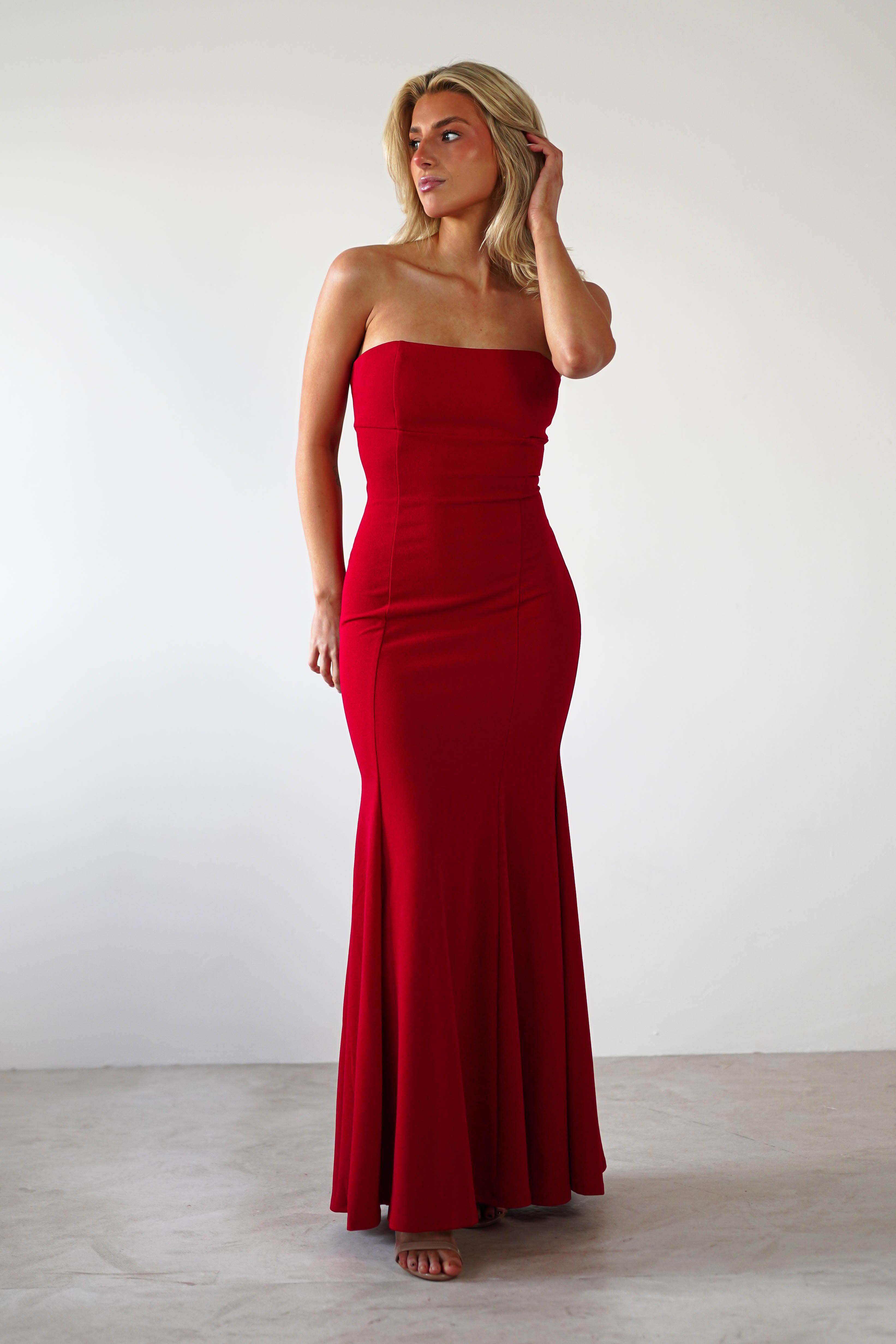 A woman stands in a strapless evening gown, the red fabric fitted at the top and elegantly flaring into a subtle mermaid silhouette. Her long, blonde hair cascades as she poses with one hand lightly touching it against a plain, light-colored background—a picture-perfect moment for formal events.