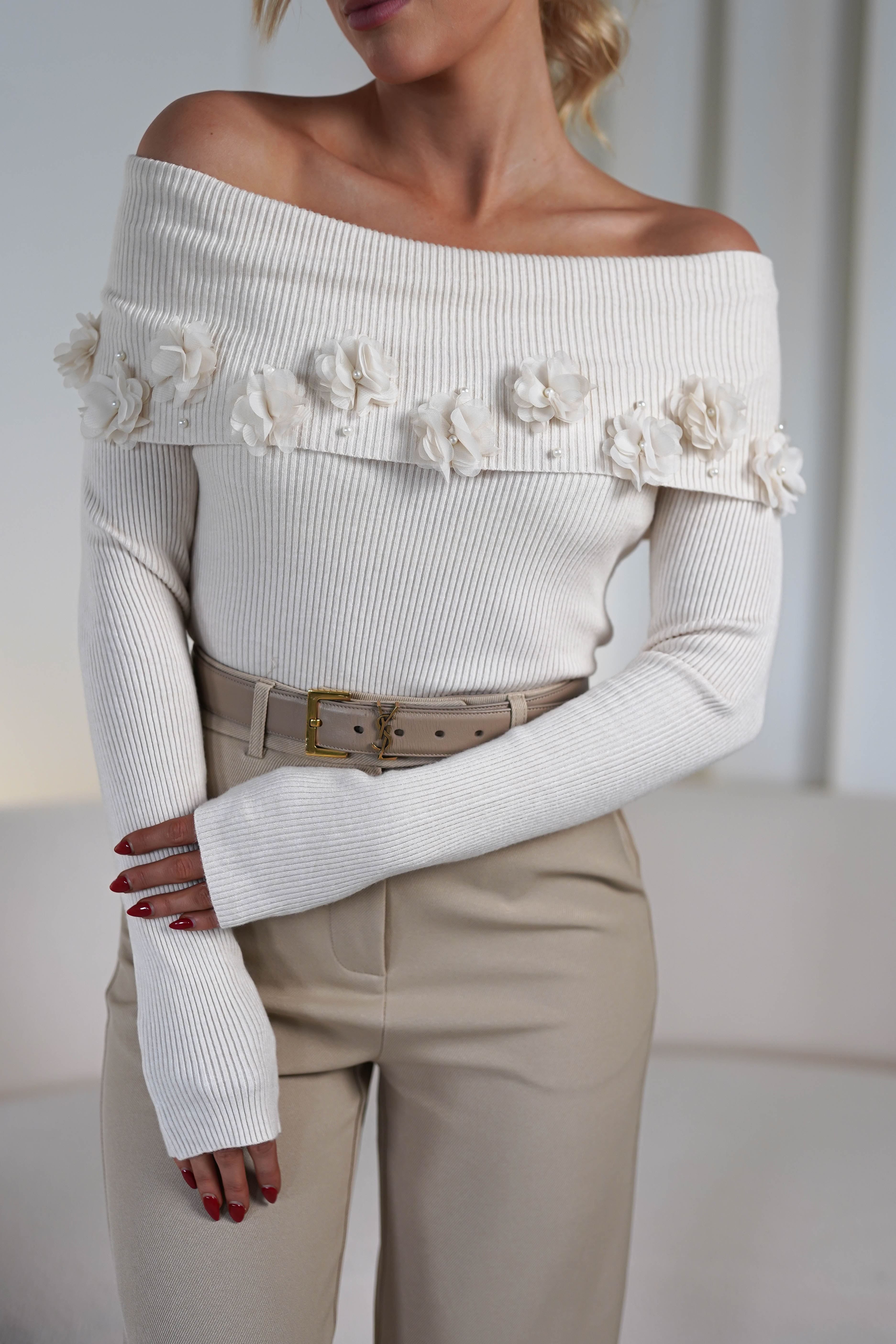 The woman dons an off-the-shoulder white sweater adorned with flower embellishments, paired gracefully with beige pants and a belt. She stands against a soft, light background in a relaxed posture, hands gently resting on her lap.