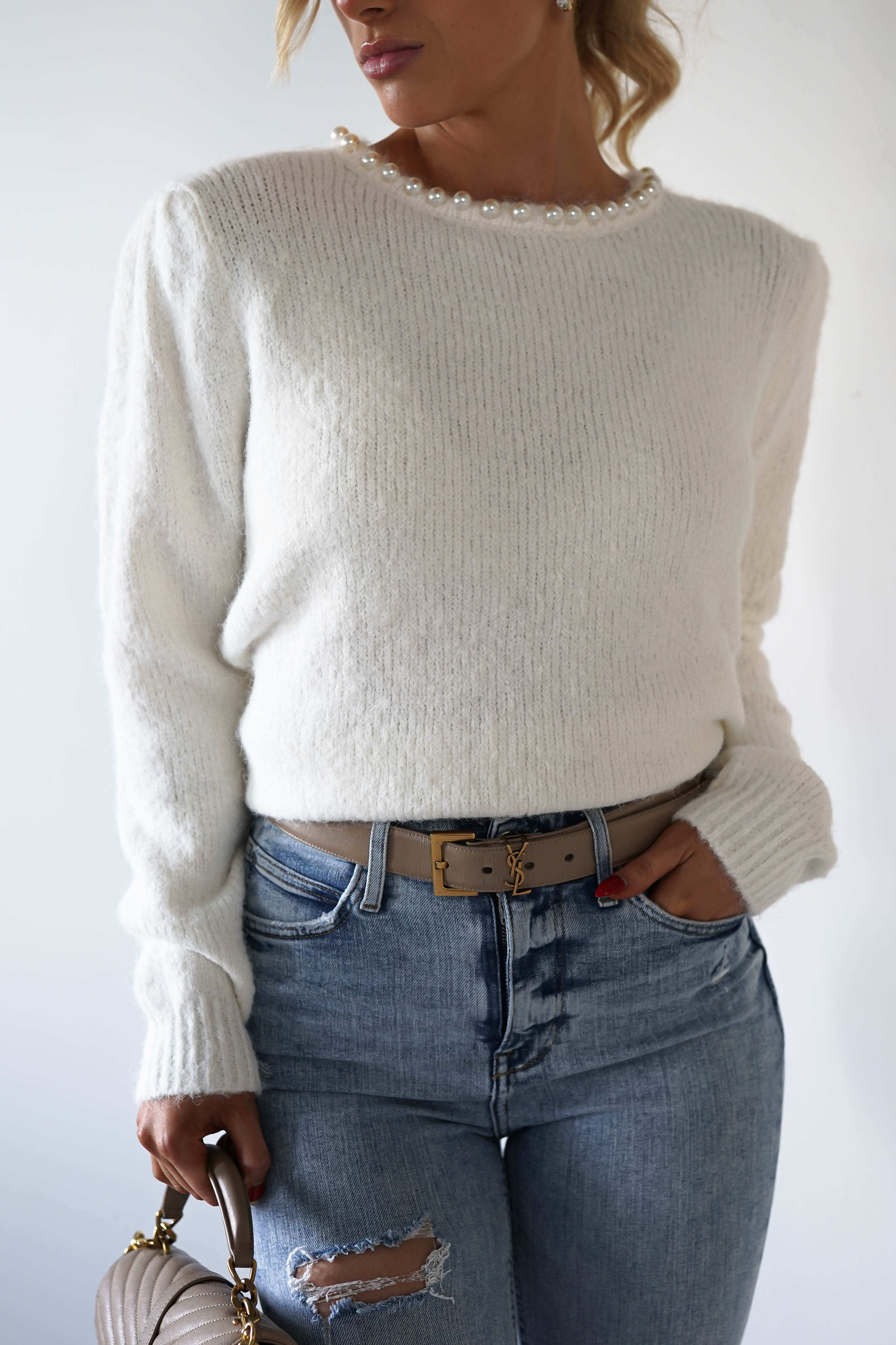 A person wearing a super soft jumper with pearl embellishments around the neckline and blue jeans. The right hand is in the jeans pocket, showcasing a belt with a gold buckle. A tan bag with brown handles hangs from the left hand.