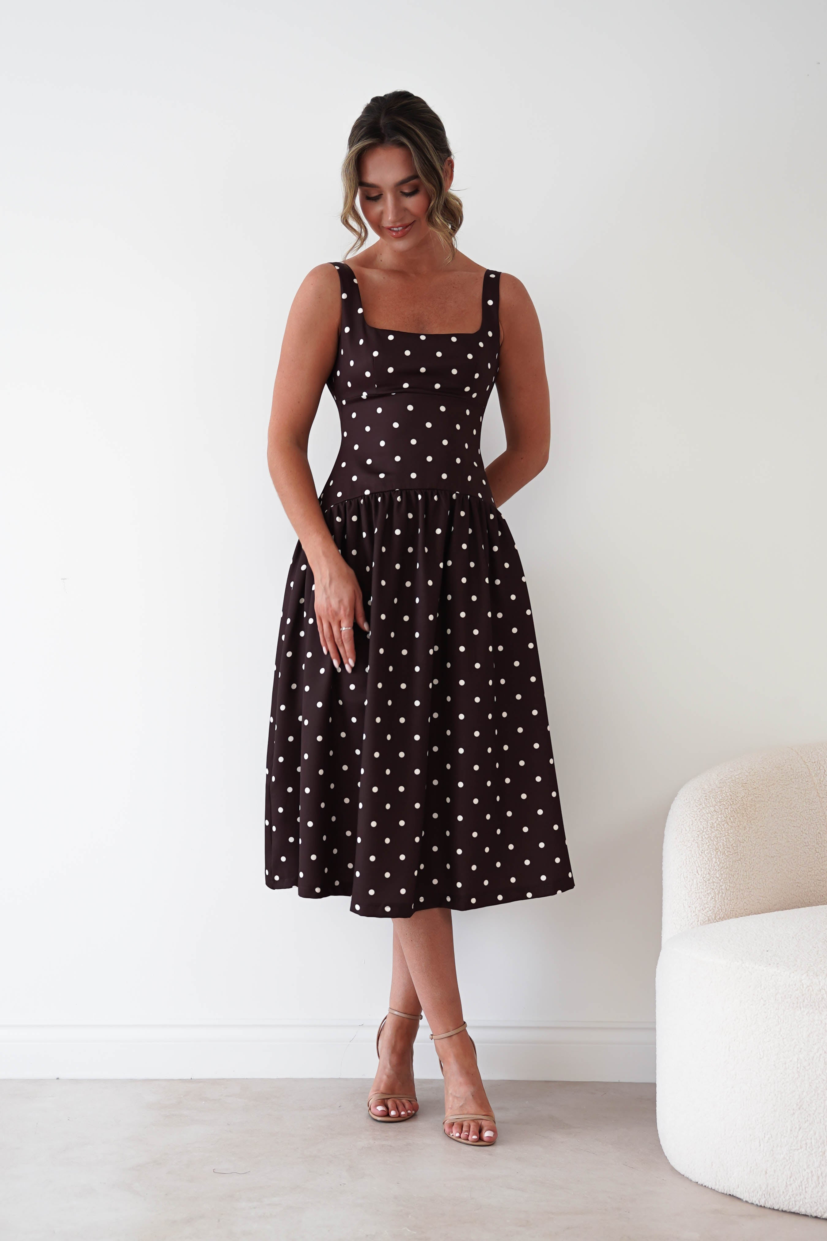 A woman modeling a sleeveless black midi dress with white polka dots, standing in a minimalist room with a white wall and a light-colored chair.