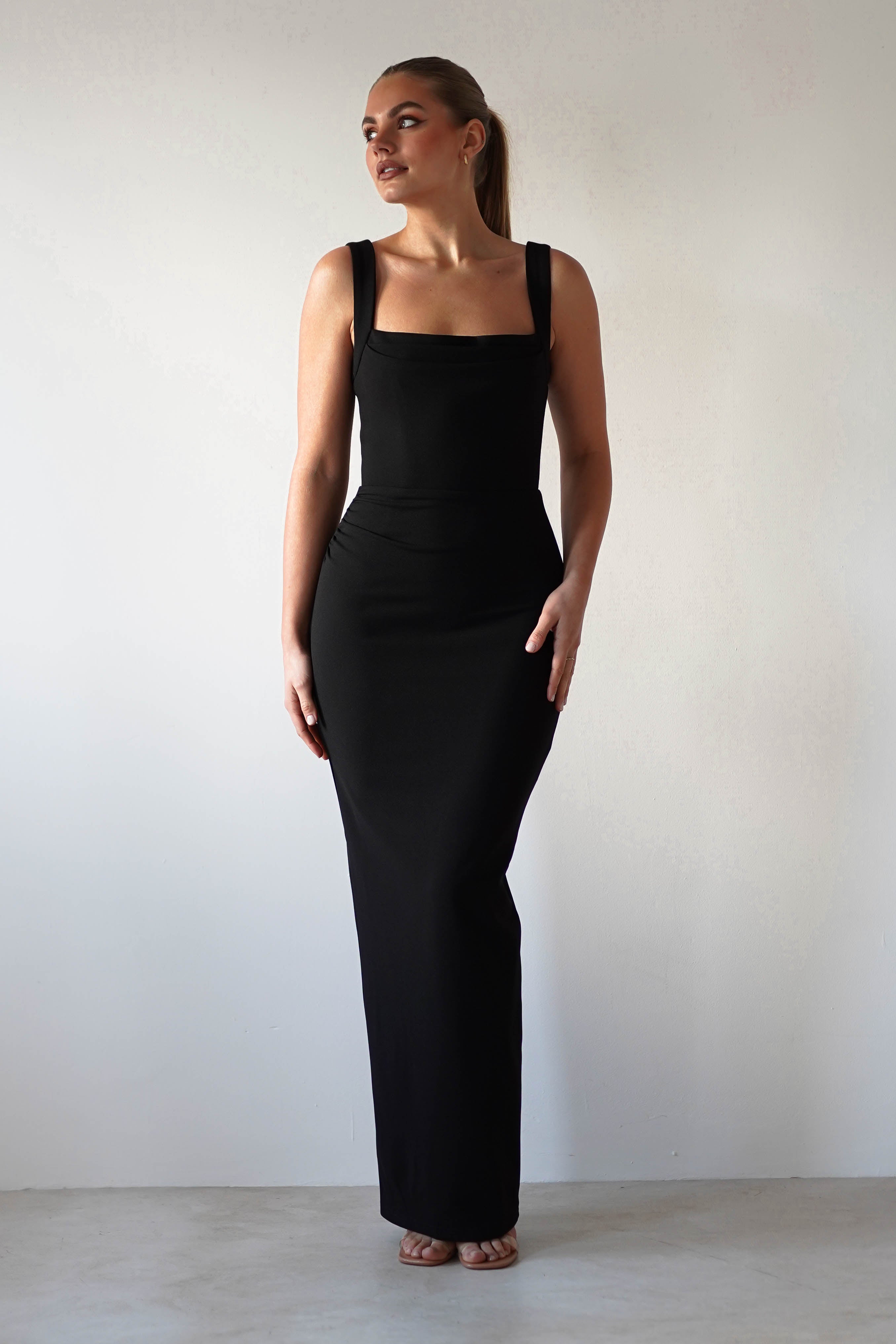 A woman poses against a white background in a glamorous, form-fitting, sleeveless square neck dress. Her hair is pulled back as she looks to the side with a neutral expression, standing elegantly on light-colored flooring.