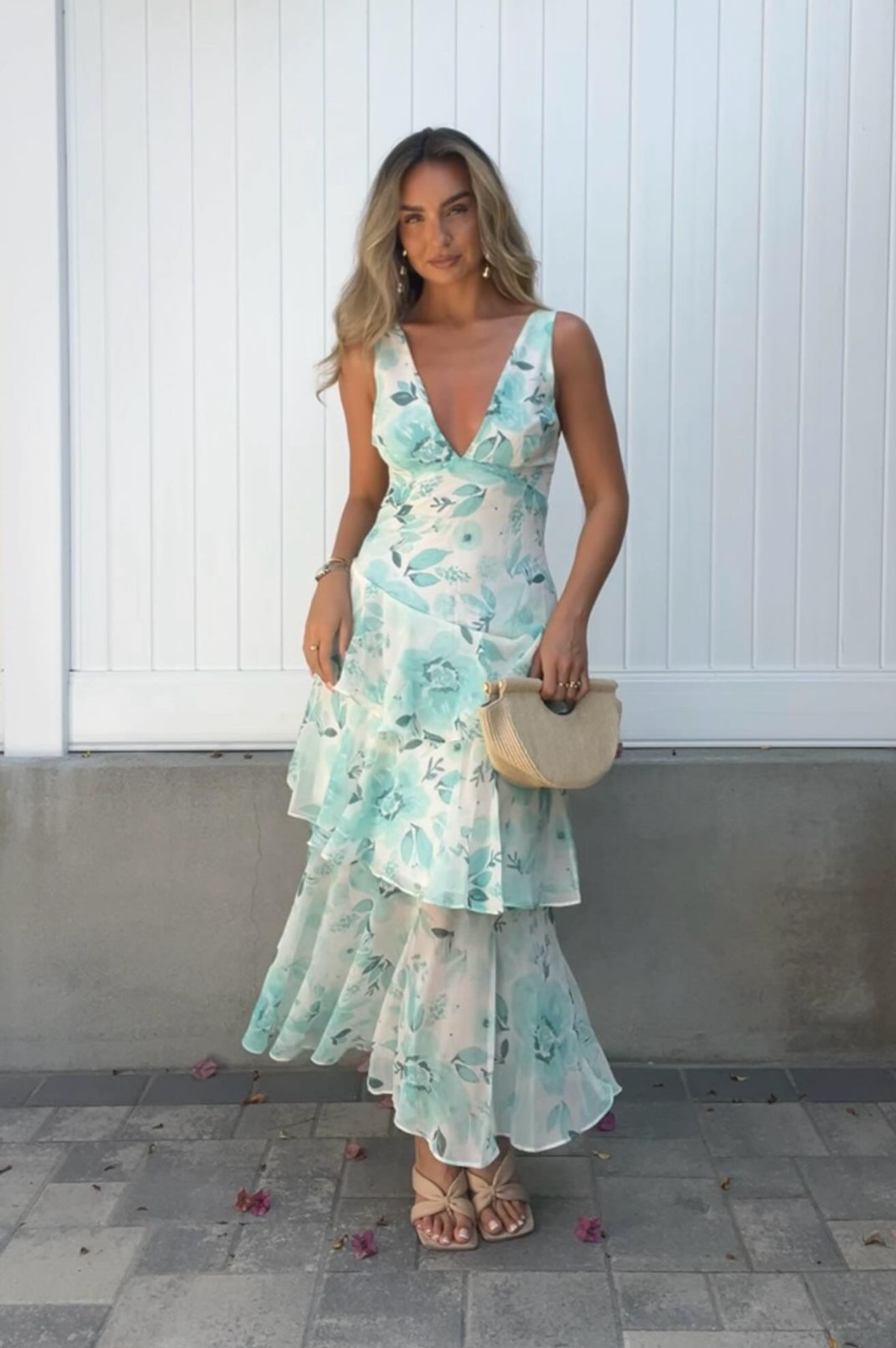 A woman poses on a paved area in front of a white paneled wall, wearing a layered, sleeveless floral dress with a deep neckline, reminiscent of a stylish wedding guest dress. She holds a round woven bag, her long hair loose and flowing as she completes the look with beige sandals.