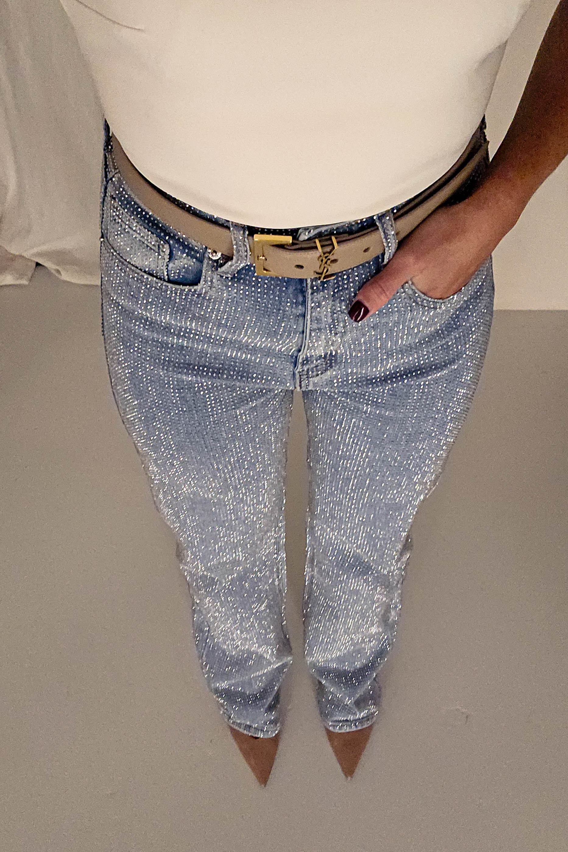 A person is standing, wearing a white top and glittery blue jeans with rhinestone embellishment and a black belt. Their left hand is in a pocket, complementing the pointed nude heels theyre sporting. The plain, light-colored background enhances the outfits allure.