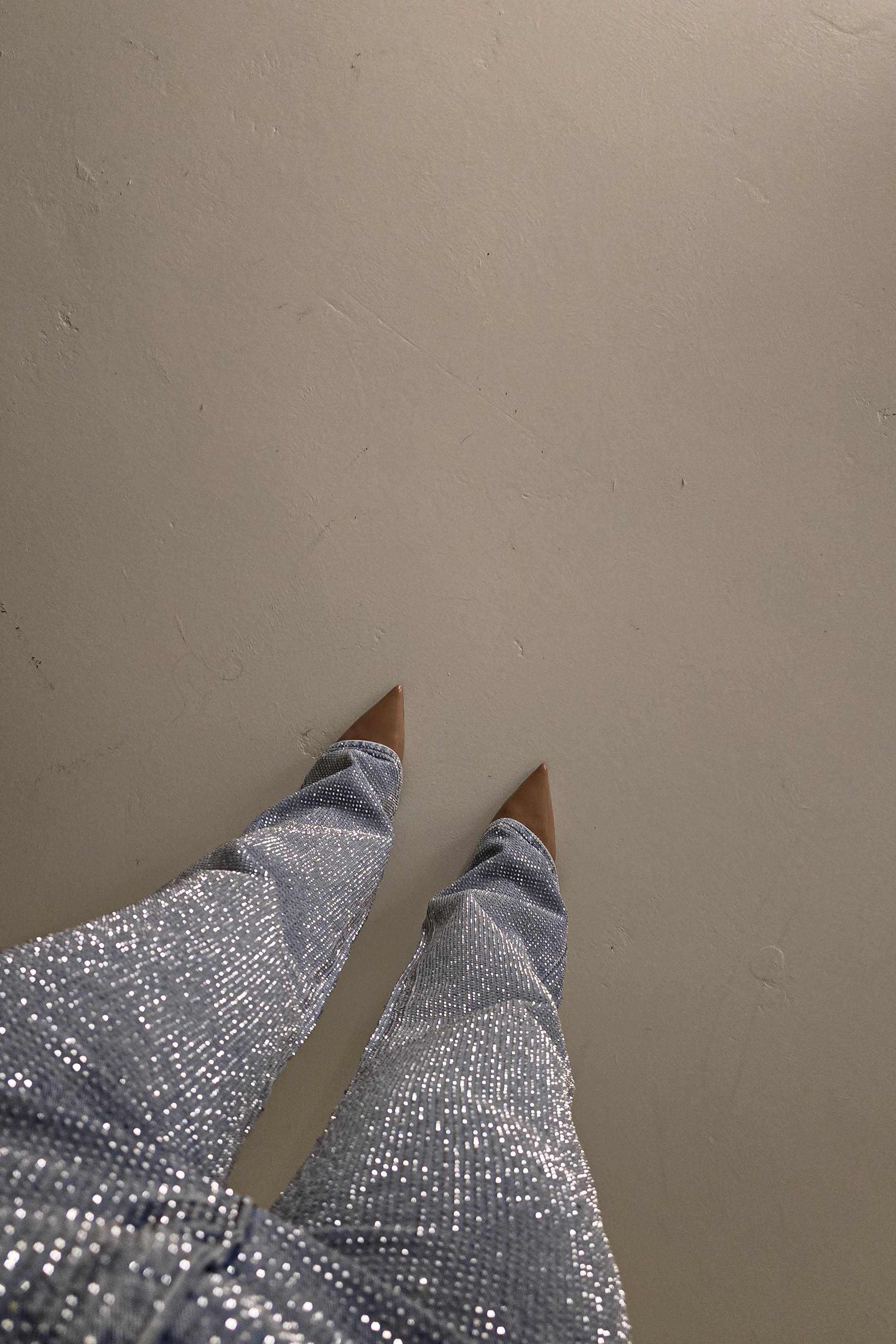 A person wearing sparkly silver pants with rhinestone embellishments and pointed brown shoes is standing on a plain, light-colored floor. The photo, taken from above, reveals only the lower part of the outfit.