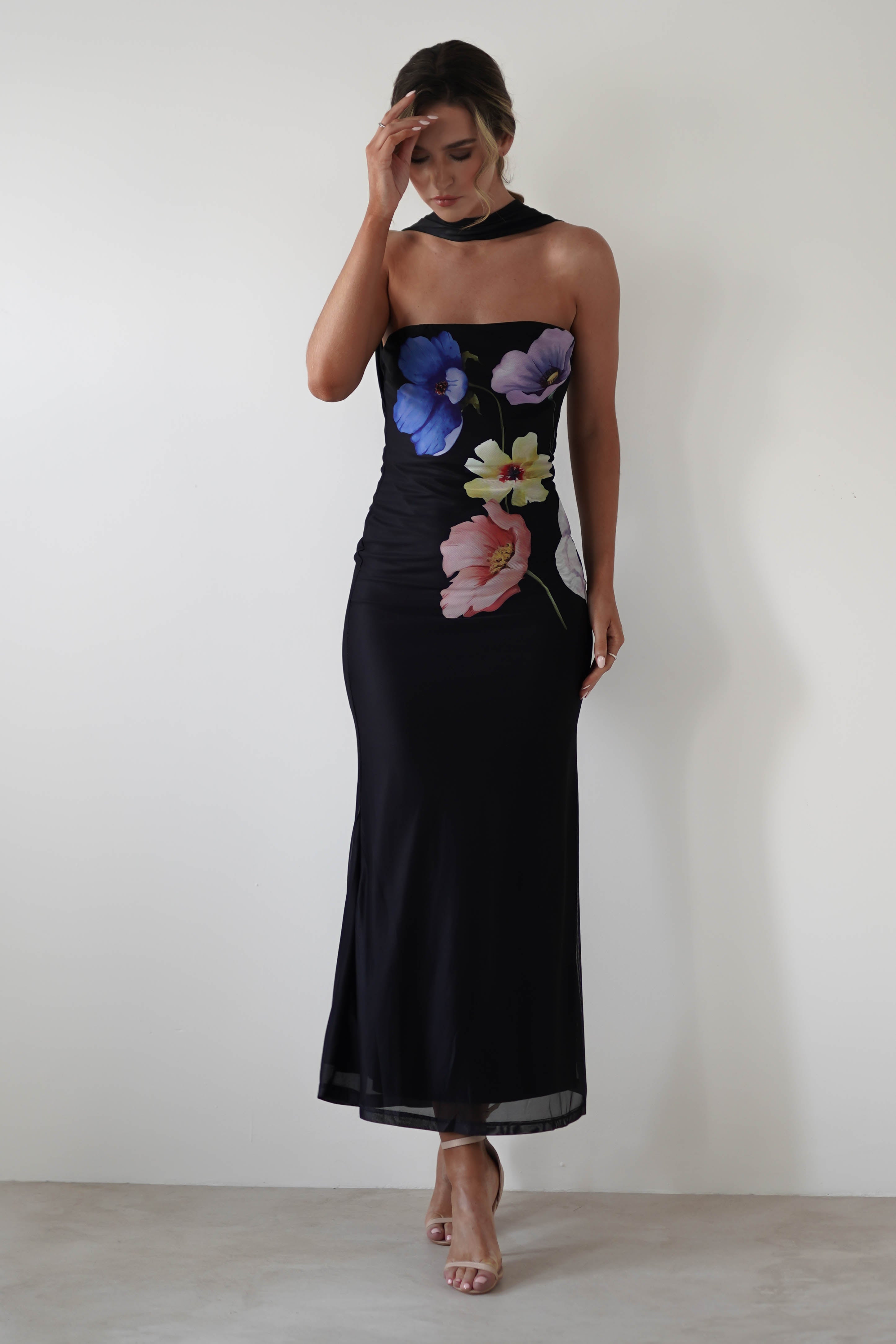 Woman in a strapless, floor-length black maxi gown with large colorful floral designs stands against a plain white background. The stretchy material gracefully hugs her figure as one hand gently touches her head, the other resting by her side.