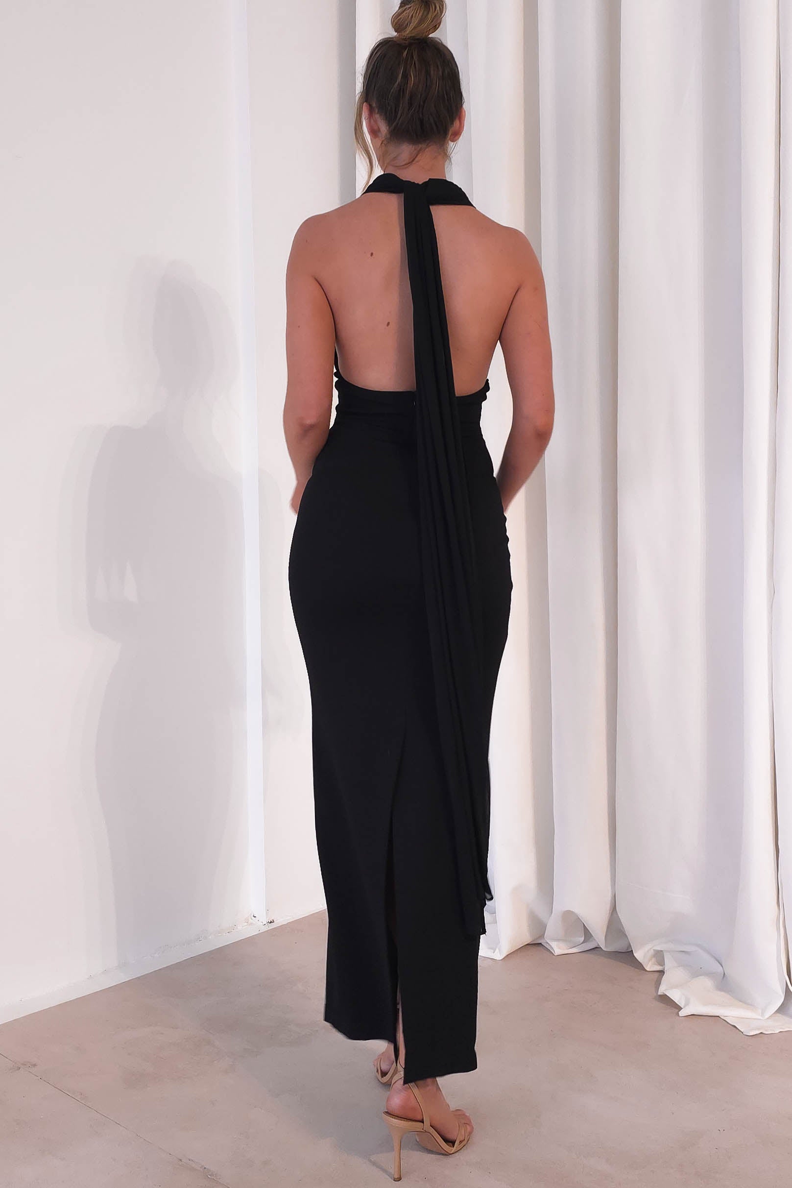 In a minimalist room with light-colored curtains, a person is elegantly dressed for formal events in a long, backless black maxi dress featuring a halter neck and flowing sash. Their hair is styled in a bun as they face away from the camera.