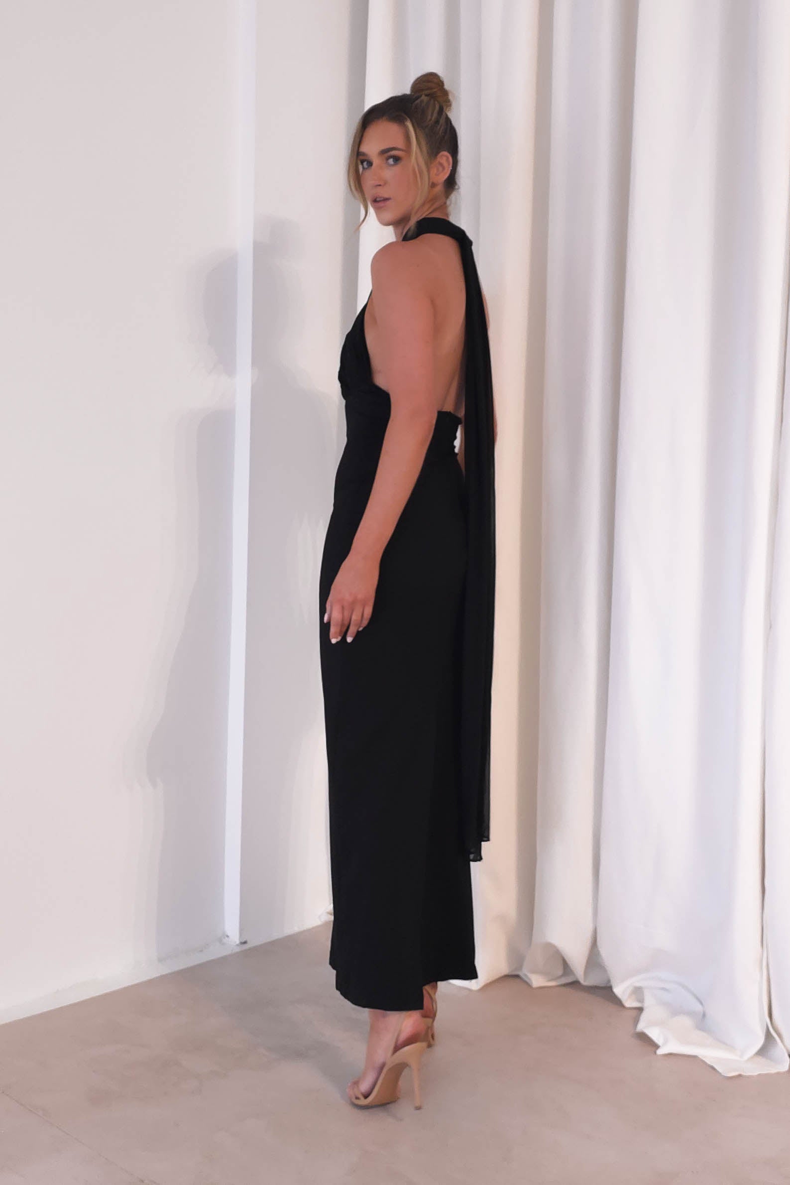 A woman in a sleek, black maxi dress with a halter neckline stands next to a white curtain. Her hair is styled in a bun, and she wears nude heels, perfectly suited for formal events. The setting appears to be indoors with minimalistic decor.