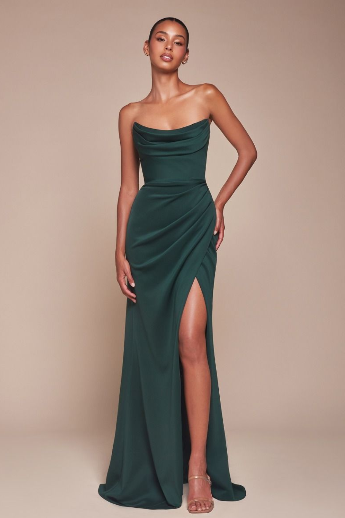 A person wearing a strapless, dark green gown with a draped bodice and thigh-high slit poses confidently. They have short hair and subtle makeup, perfect for a formal occasion. The background is a neutral beige tone.