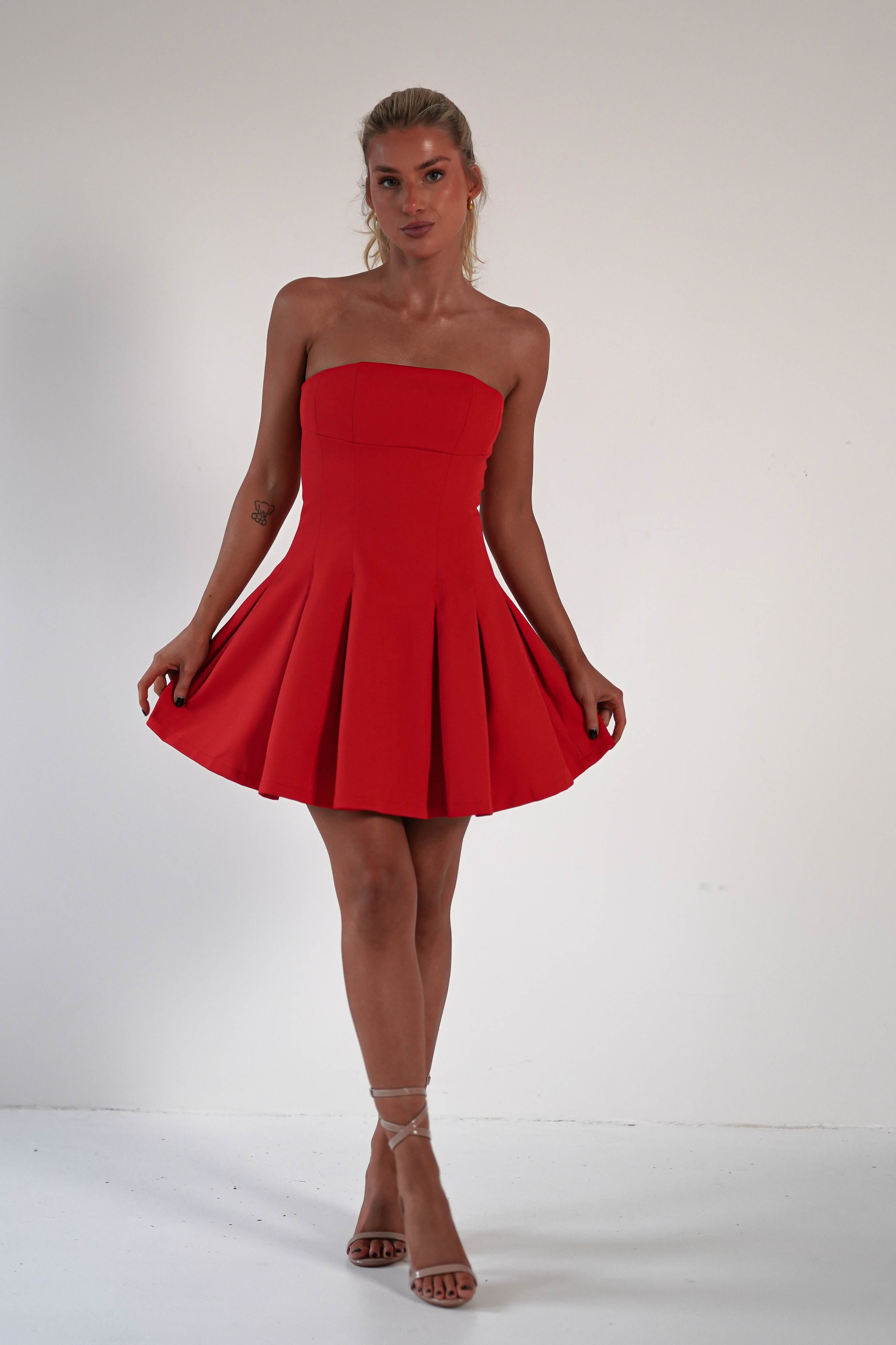 Person in a strapless red mini dress with a pleated skirt, perfect as a wedding guest dress, standing against a plain white background. They hold the sides of the skirt, showcasing its shape. They wear strappy high-heeled sandals.