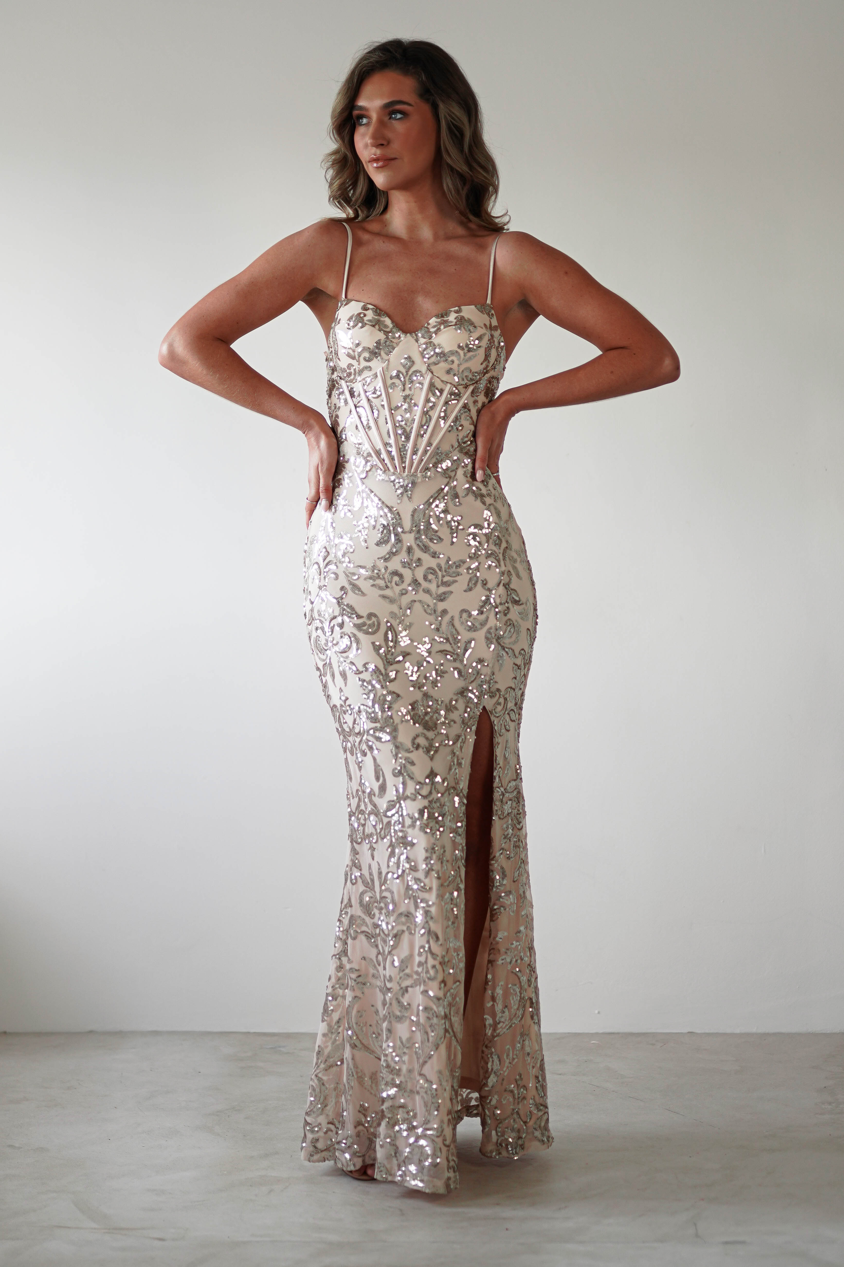 A person in a floor-length Champagne gown with intricate sequin embroidery and a thigh-high slit stands against a plain background. The dress features thin straps and a structured bodice as they pose with hands on hips.