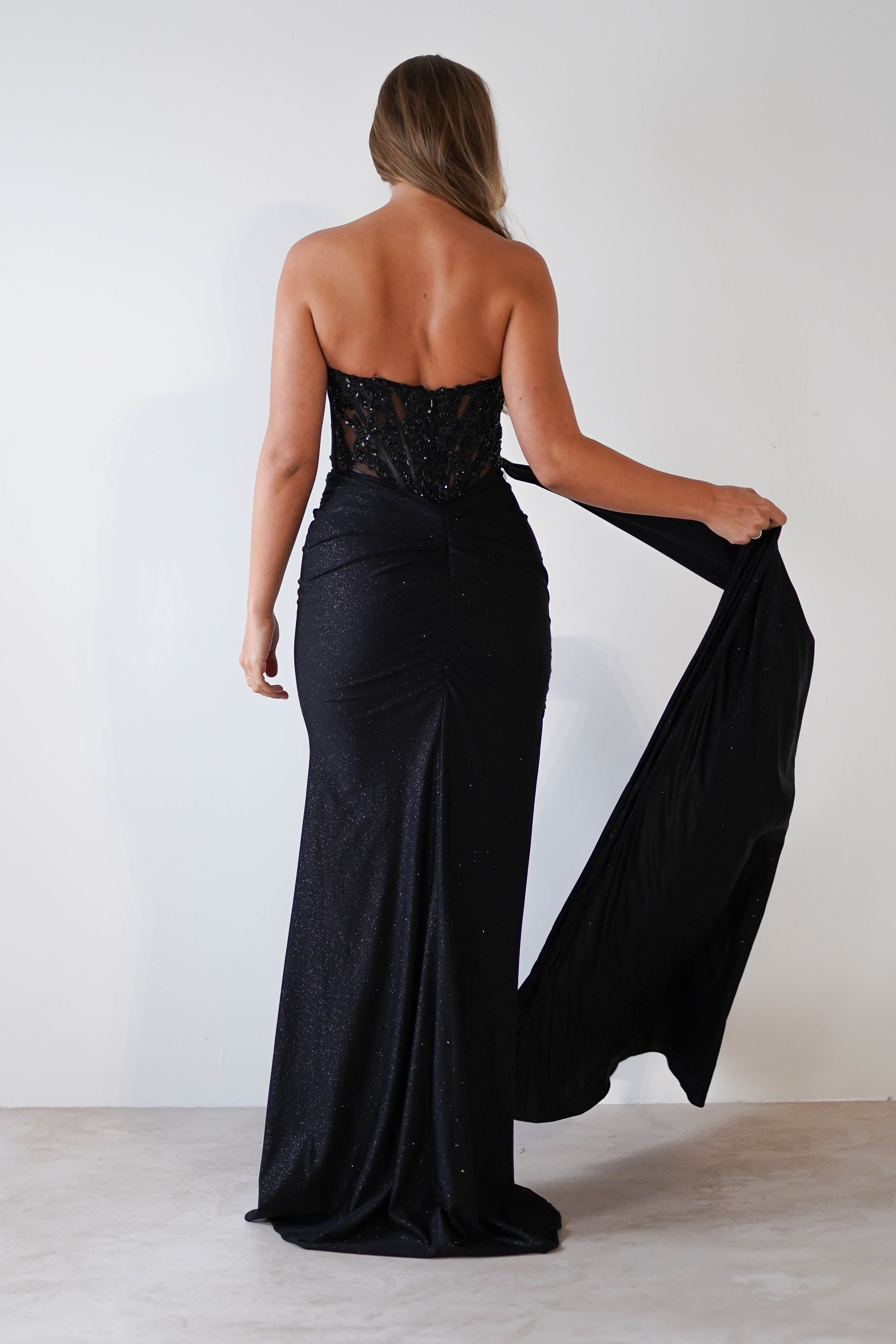 A woman with long hair is wearing a stunning black glitter gown, complete with a fitted design and intricate lace detailing at the back. As she stands on a plain floor, facing away and holding the train, the gowns elegance makes it perfect for formal events.