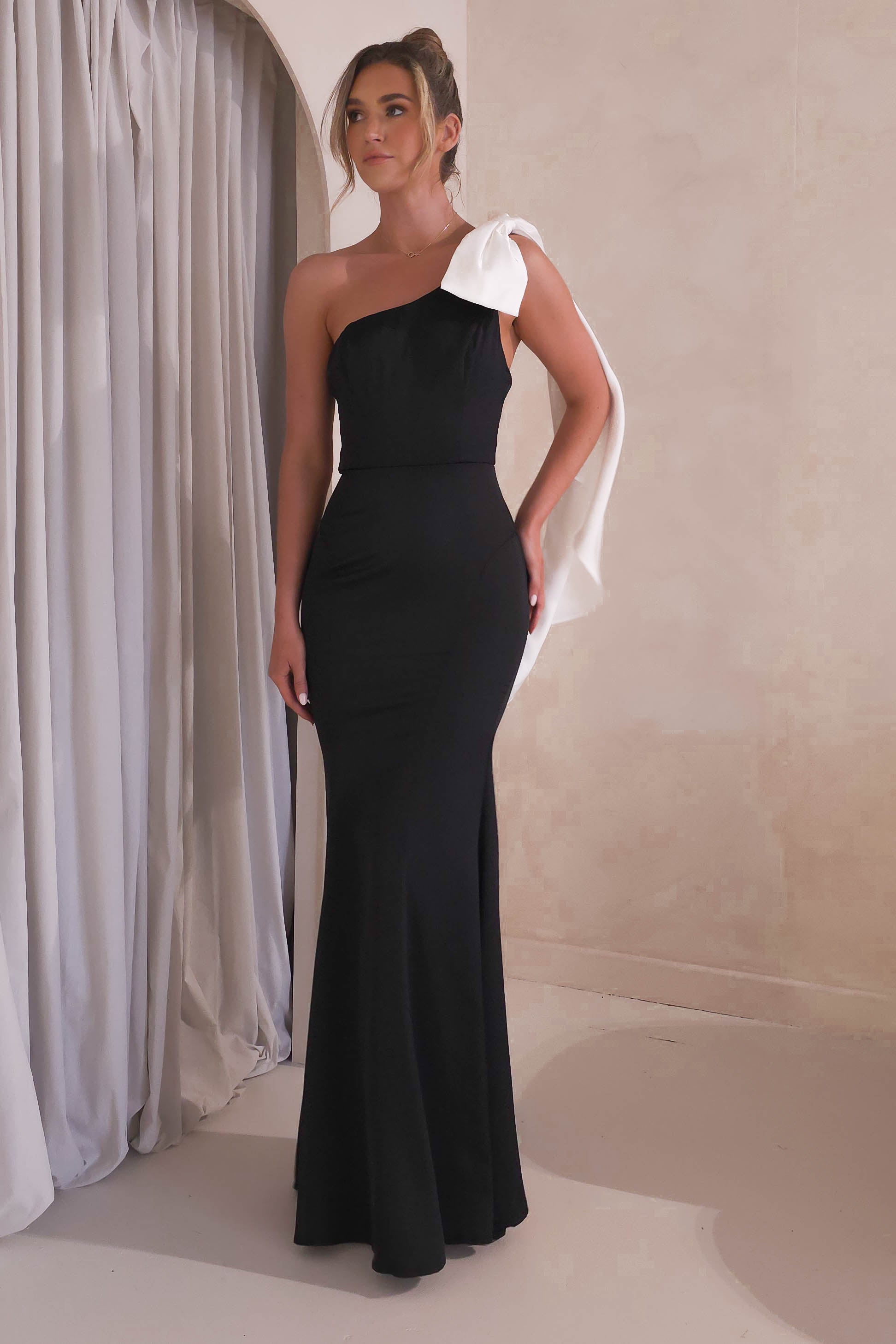 A woman poses elegantly in a sleek satin one-shoulder gown, featuring a large white bow on the shoulder. She stands gracefully before a light curtain and neutral-toned background, her hair styled up with sophistication.