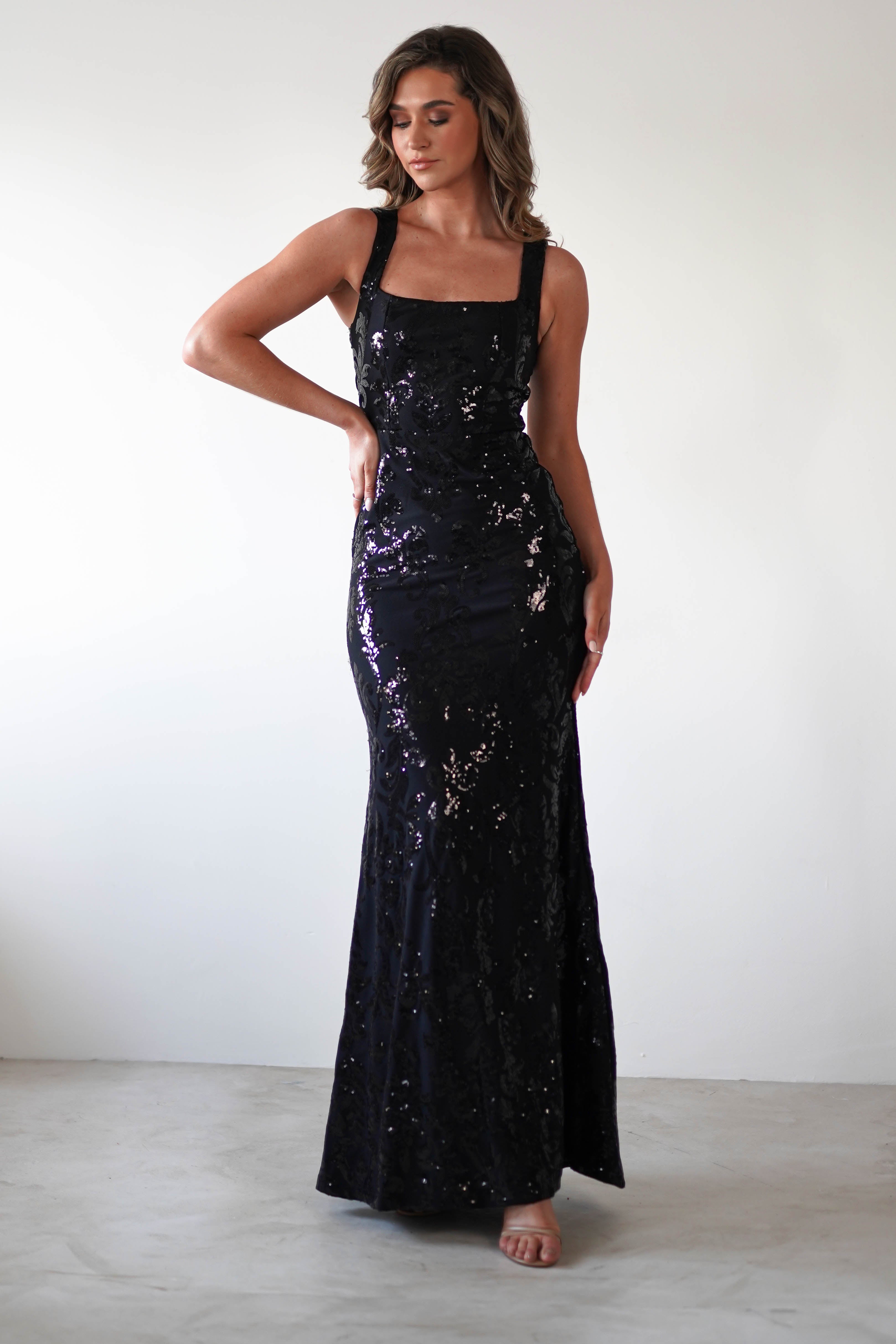 A woman in a sparkling, dark blue sequined evening gown poses against a plain white background. The black gown features a square neckline and wide shoulder straps, as she stands with one hand on her hip, exuding elegance and confidence.