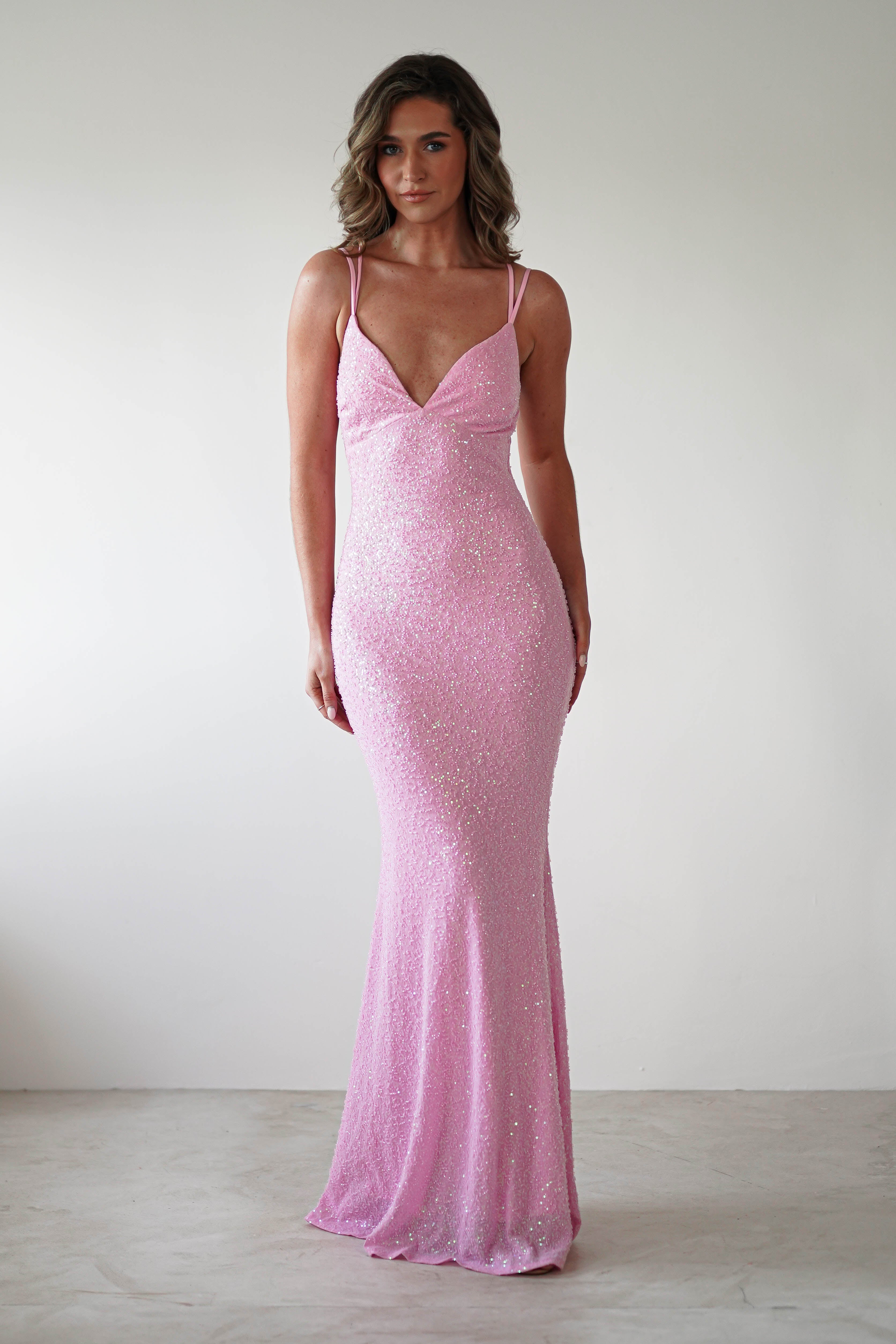 A woman graces the scene in a sequin gown perfect for special occasions, styled with thin straps and a V-neckline. Her sparkly, form-fitting pink dress contrasts elegantly against the plain white background. With wavy, shoulder-length hair and a neutral expression, she captivates effortlessly.