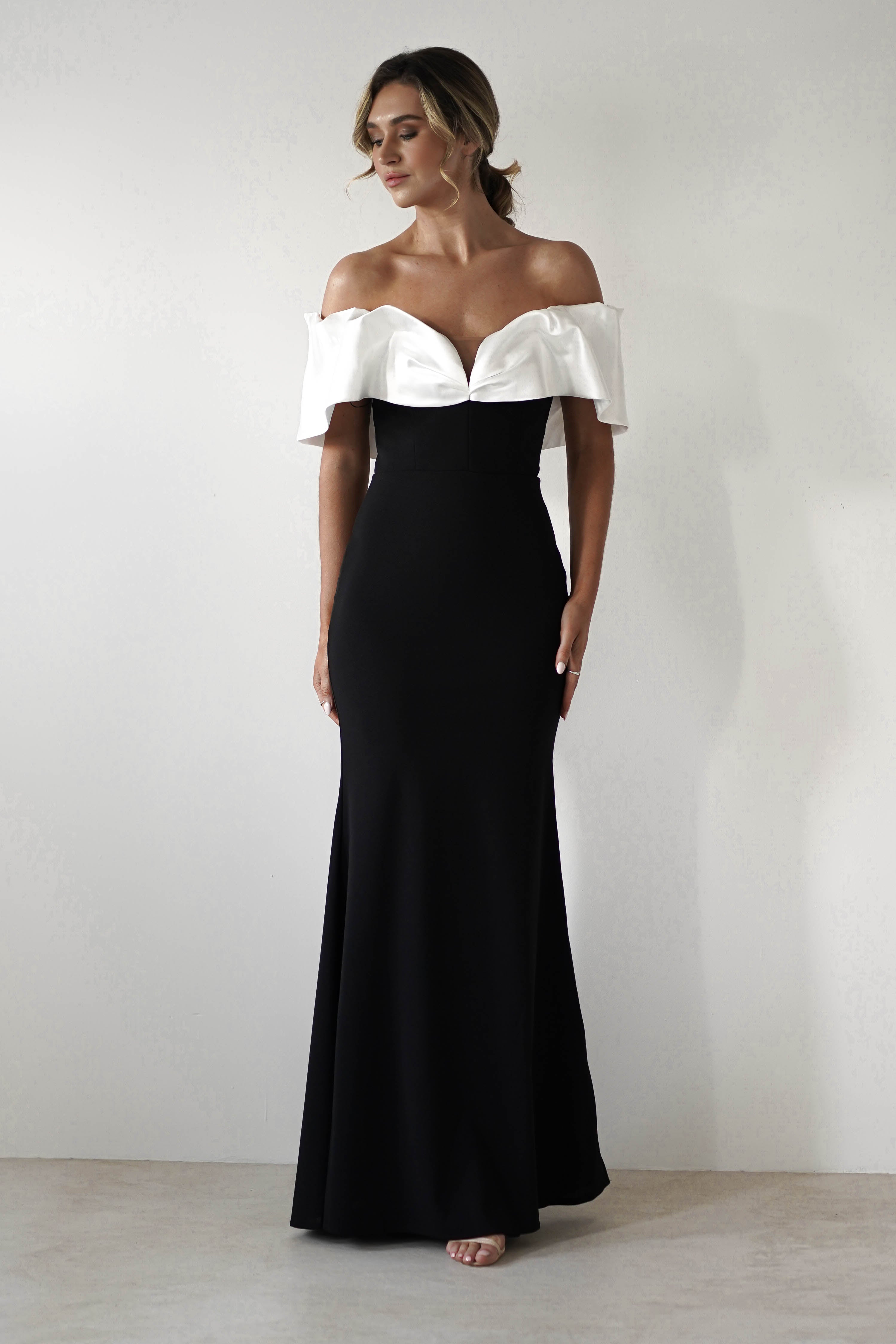 A person wearing a stunning, off-shoulder evening gown with a sweetheart neckline stands against a plain background. The black dress with a white ruffled bodice flows into a mermaid maxi style, and the individual, with styled hair, looks to the side. Perfect for formal events.