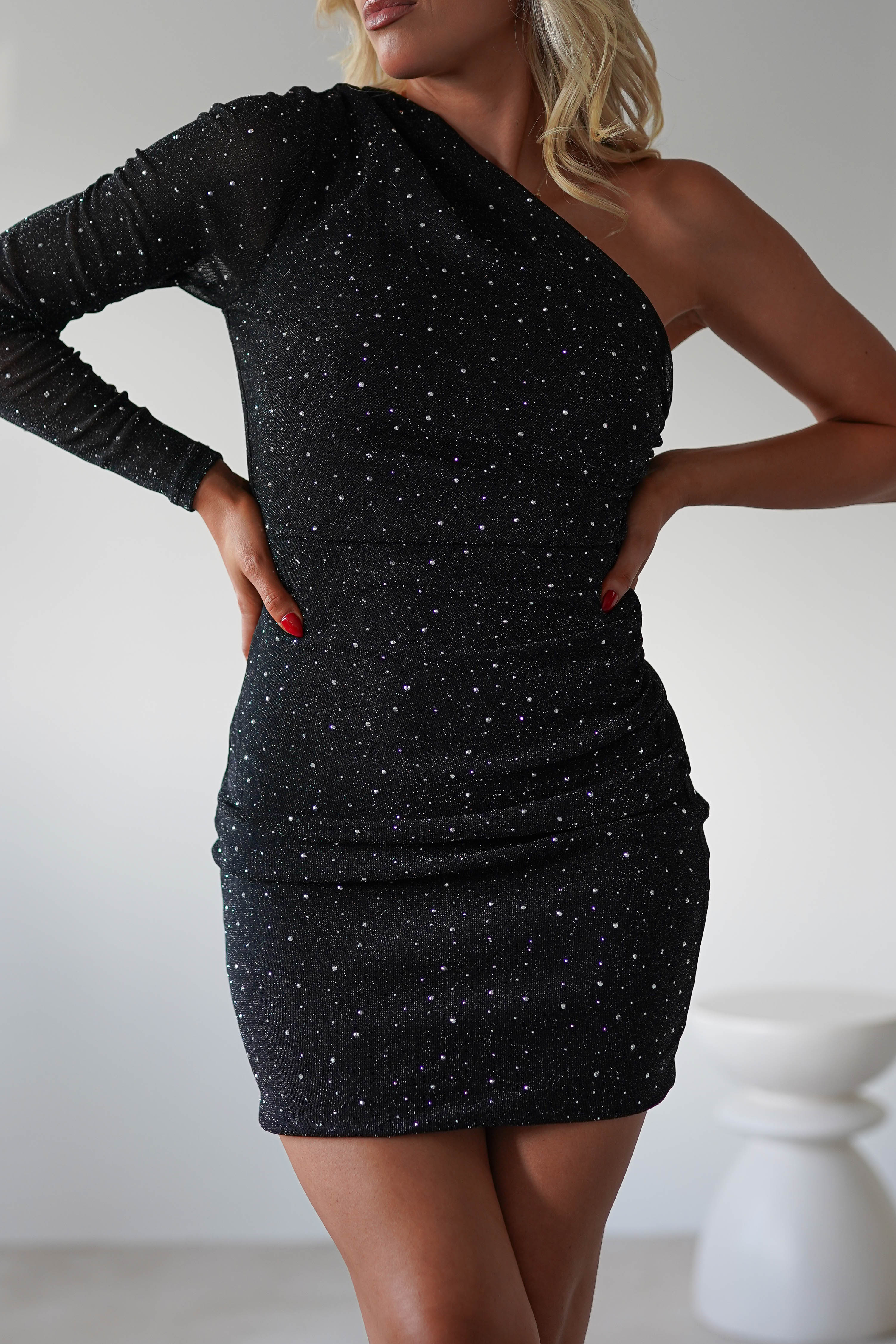 A person posing in a sparkly, one-shoulder black party mini dress adorned with diamante detail and featuring one long sleeve. Their hands rest confidently on their hips against a plain, minimalistic background.